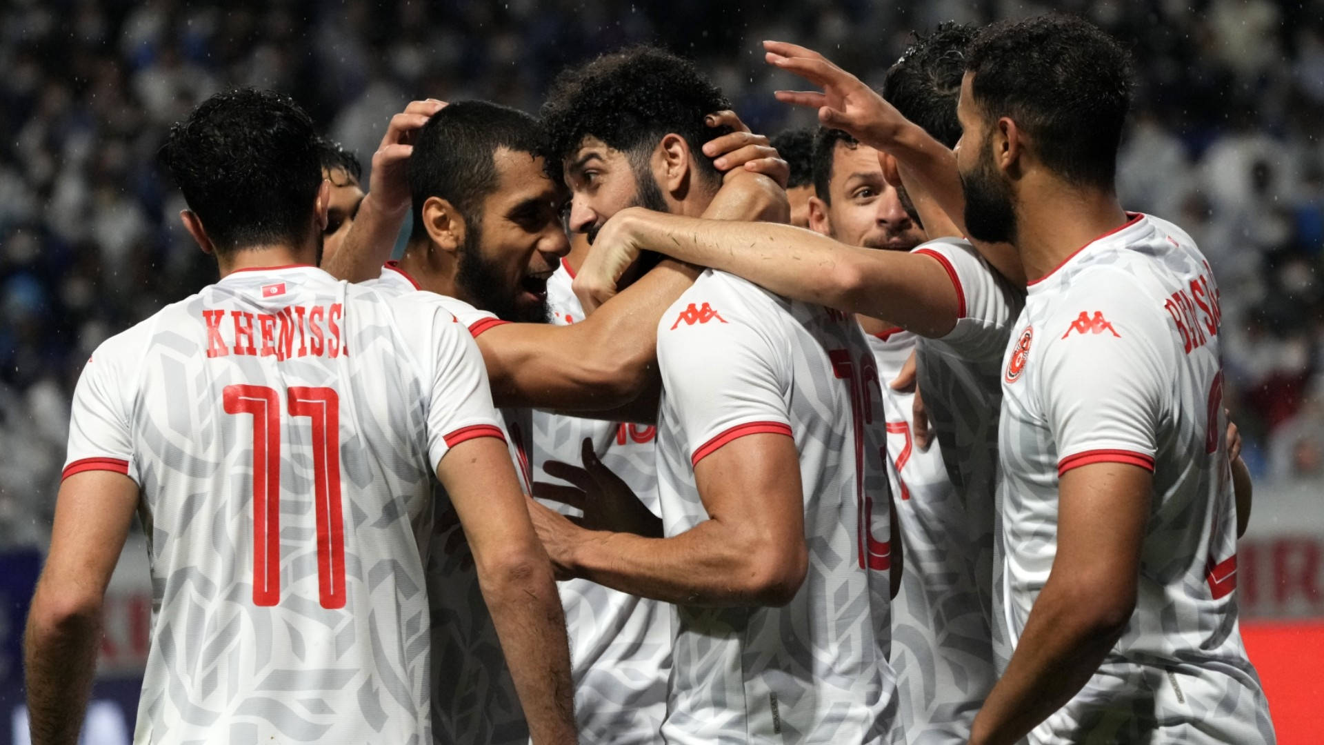 Tunisia 桌布 壁紙 — 身穿白色球衣帶紅色點綴的足球運動員在球場燈光下緊緊擁抱慶祝進球。