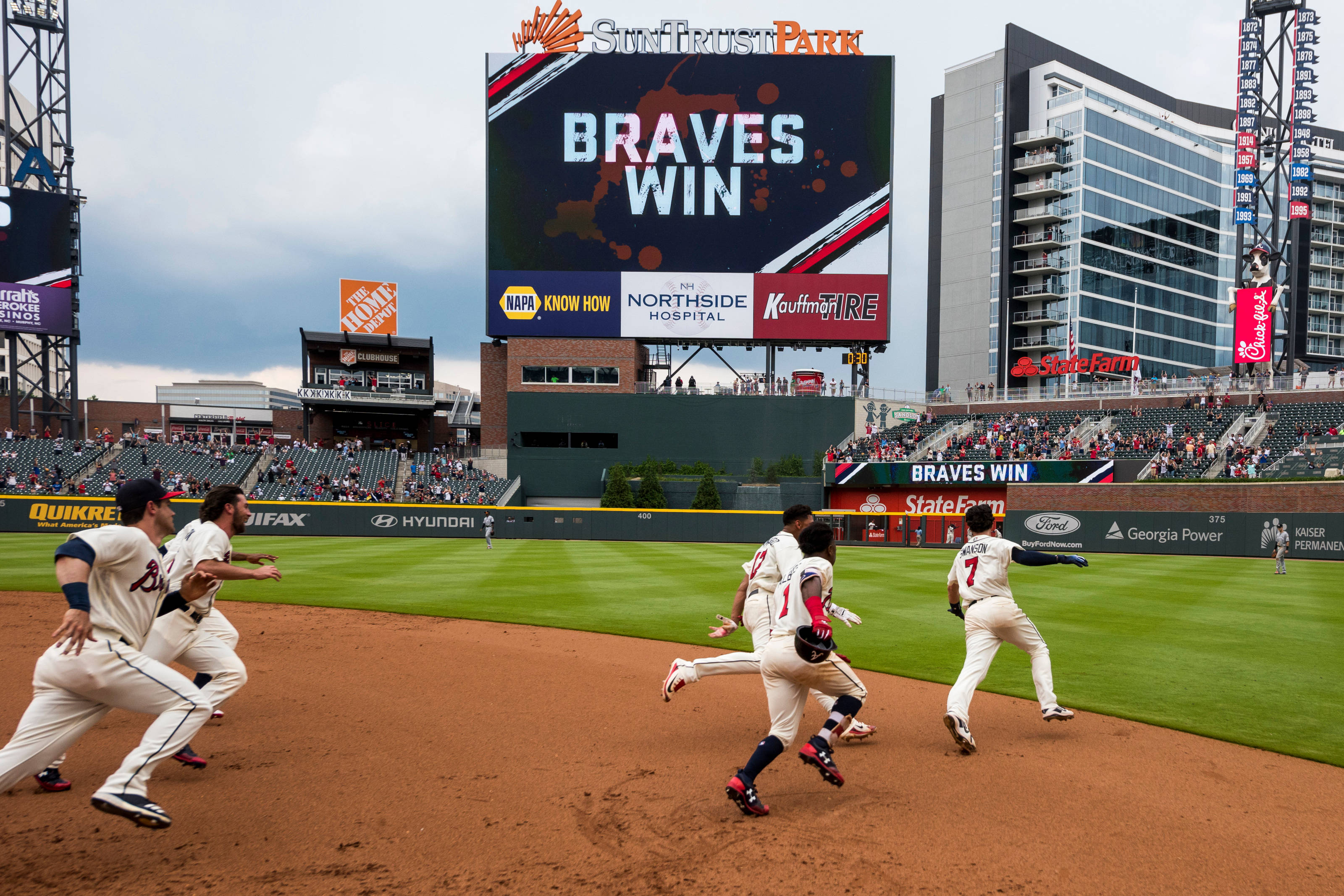 Braves 桌布 壁紙 — 身穿白色球衣的棒球運動員在陽光明媚的球場上奔跑，巨大的記分牌在陰天天空下宣告「BRAVES WIN」。