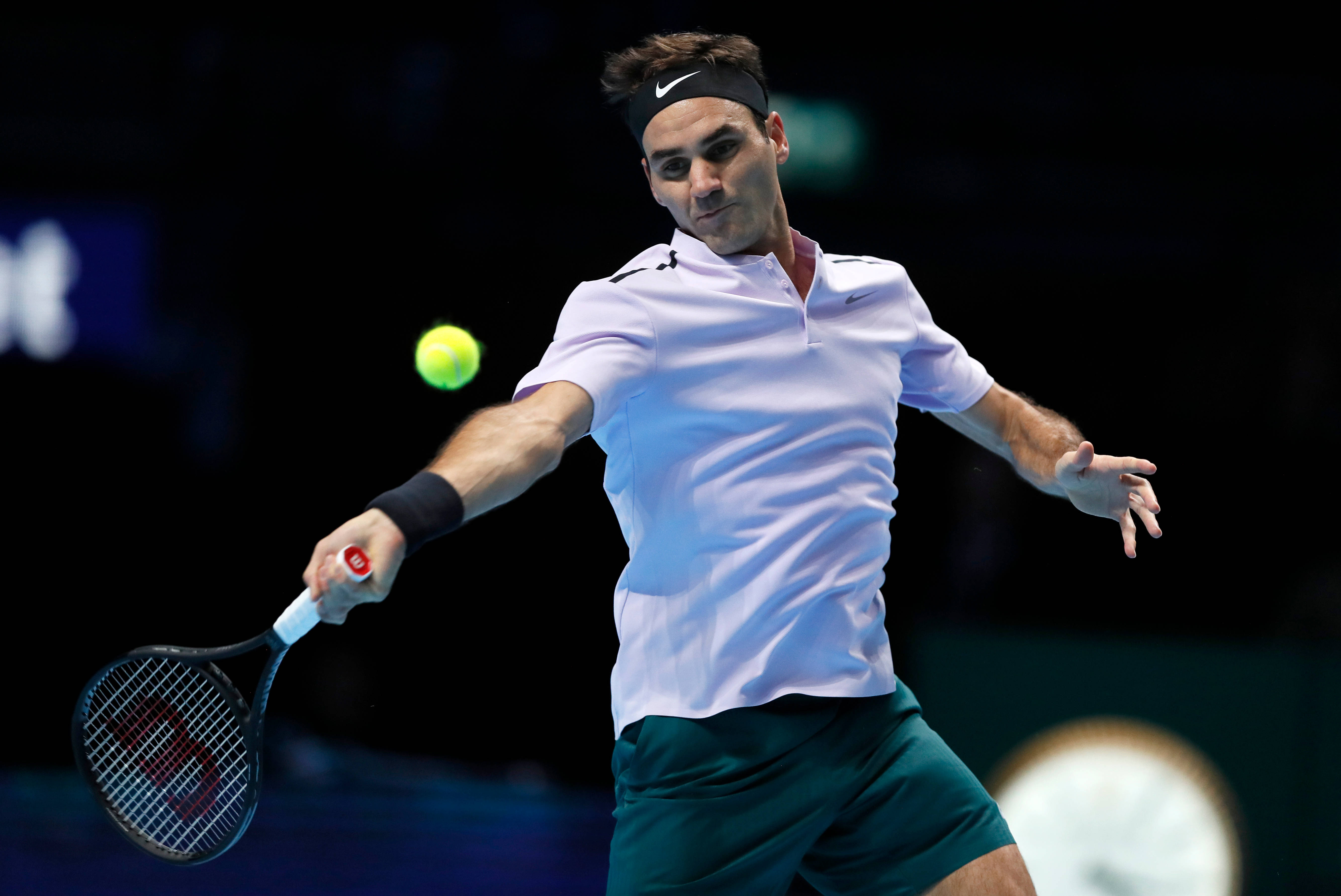 roger 桌布 壁紙 — 身穿白色襯衫和綠色短褲的網球選手在黑暗的競技場背景下揮拍擊打發光的黃色球。