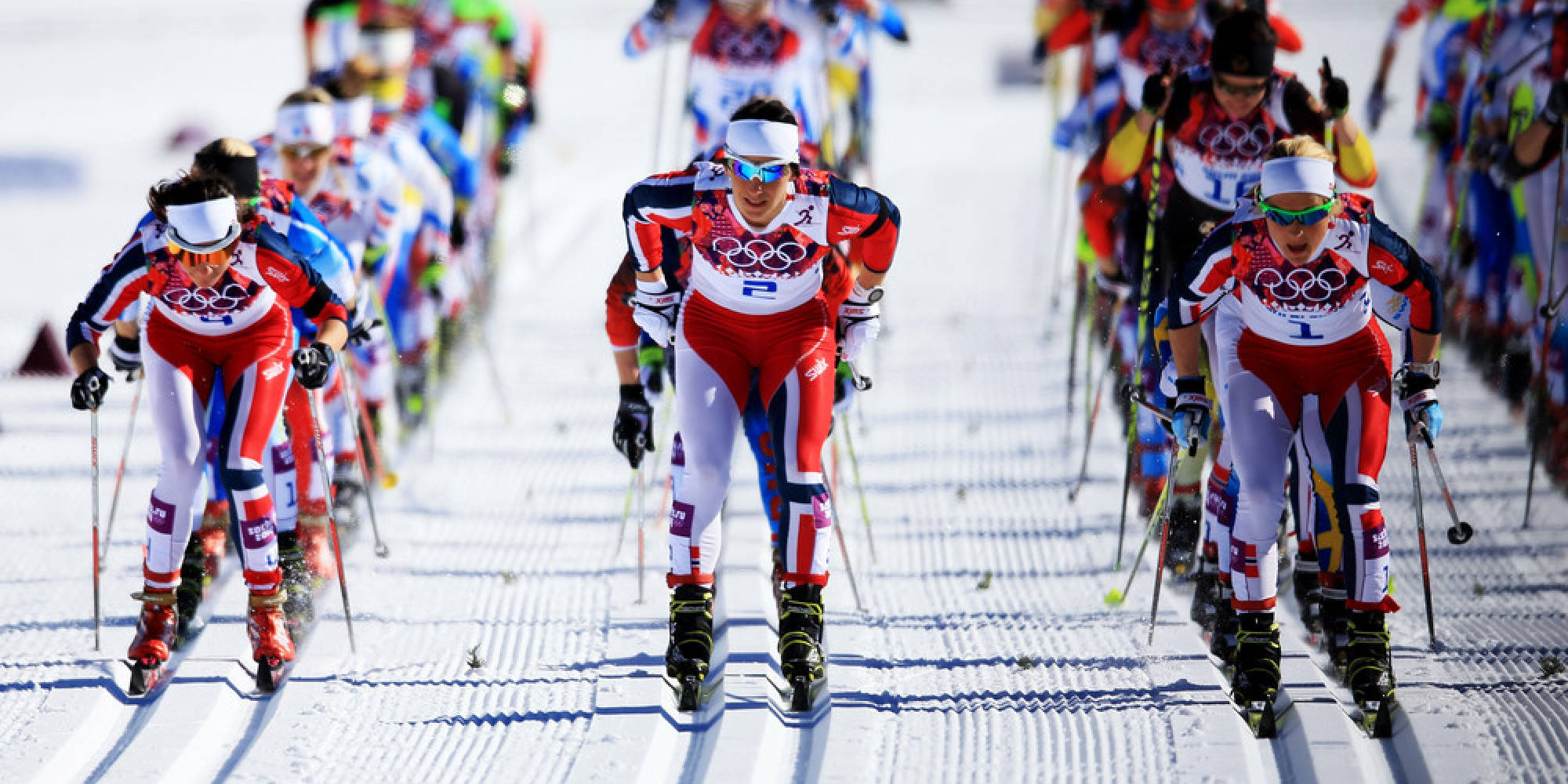 Winter Olympics 桌布 壁紙 — 身穿紅、白、藍色制服的競技越野滑雪選手在明亮的陽光下沿著修整過的雪道飛馳。
