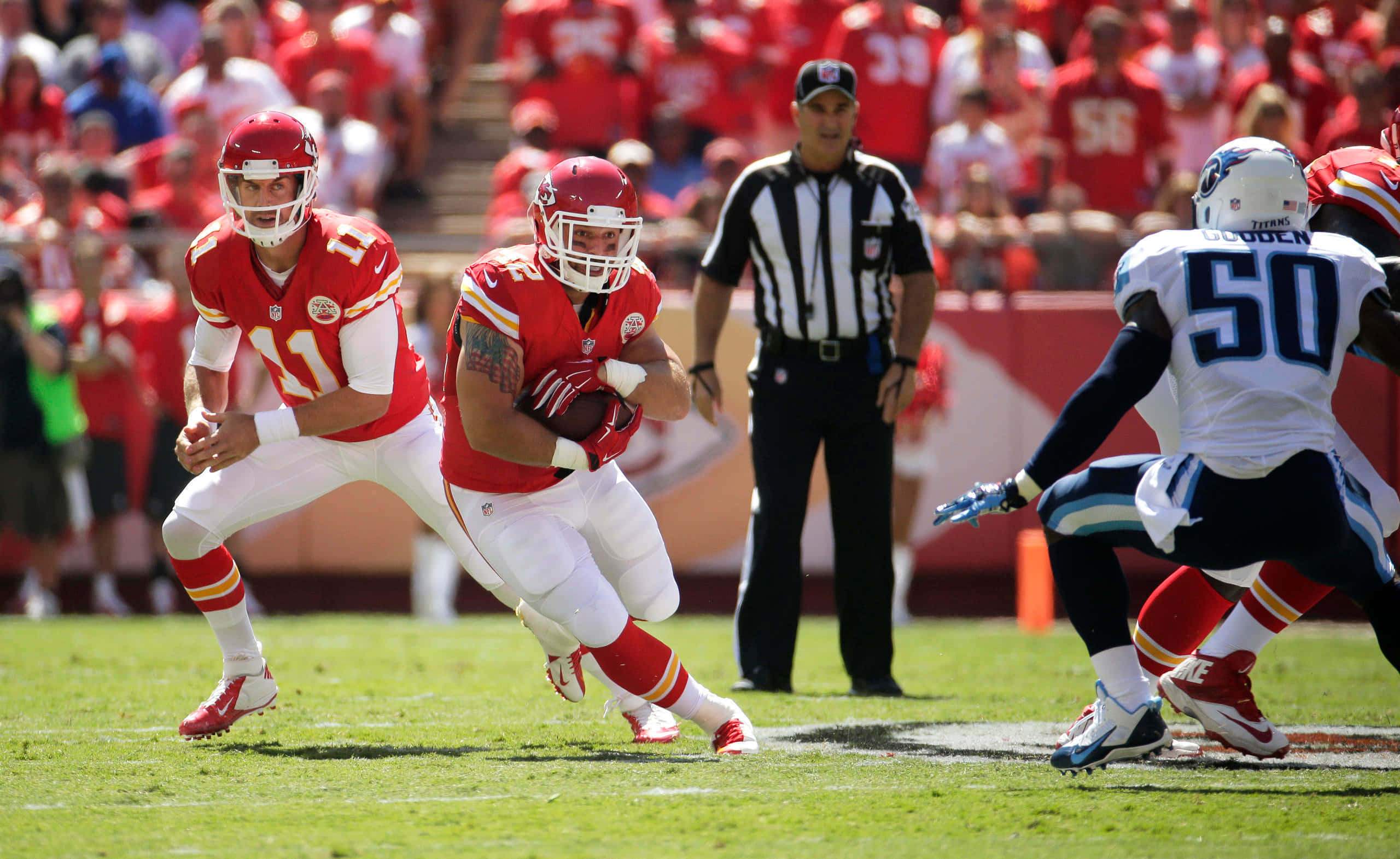 Kansas City Chiefs 4K 桌布 壁紙 — 身穿紅白球衣的美式足球員在陽光明媚的綠色球場上比賽中衝刺，背景有裁判和人群。
