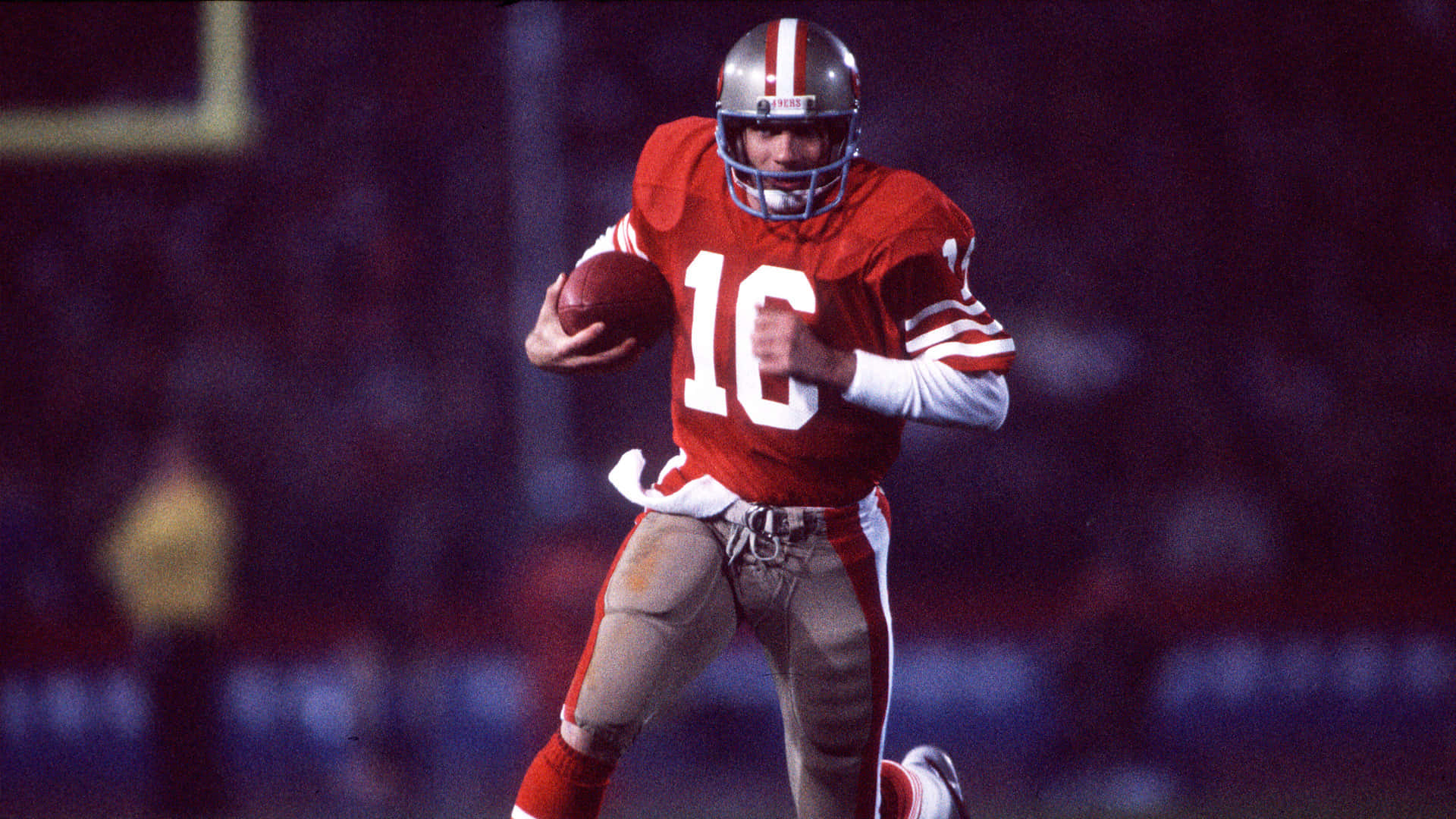 Joe Montana 桌布 壁紙 — 身穿紅色16號球衣的美式足球四分衛在夜間球場燈光下帶球奔跑。