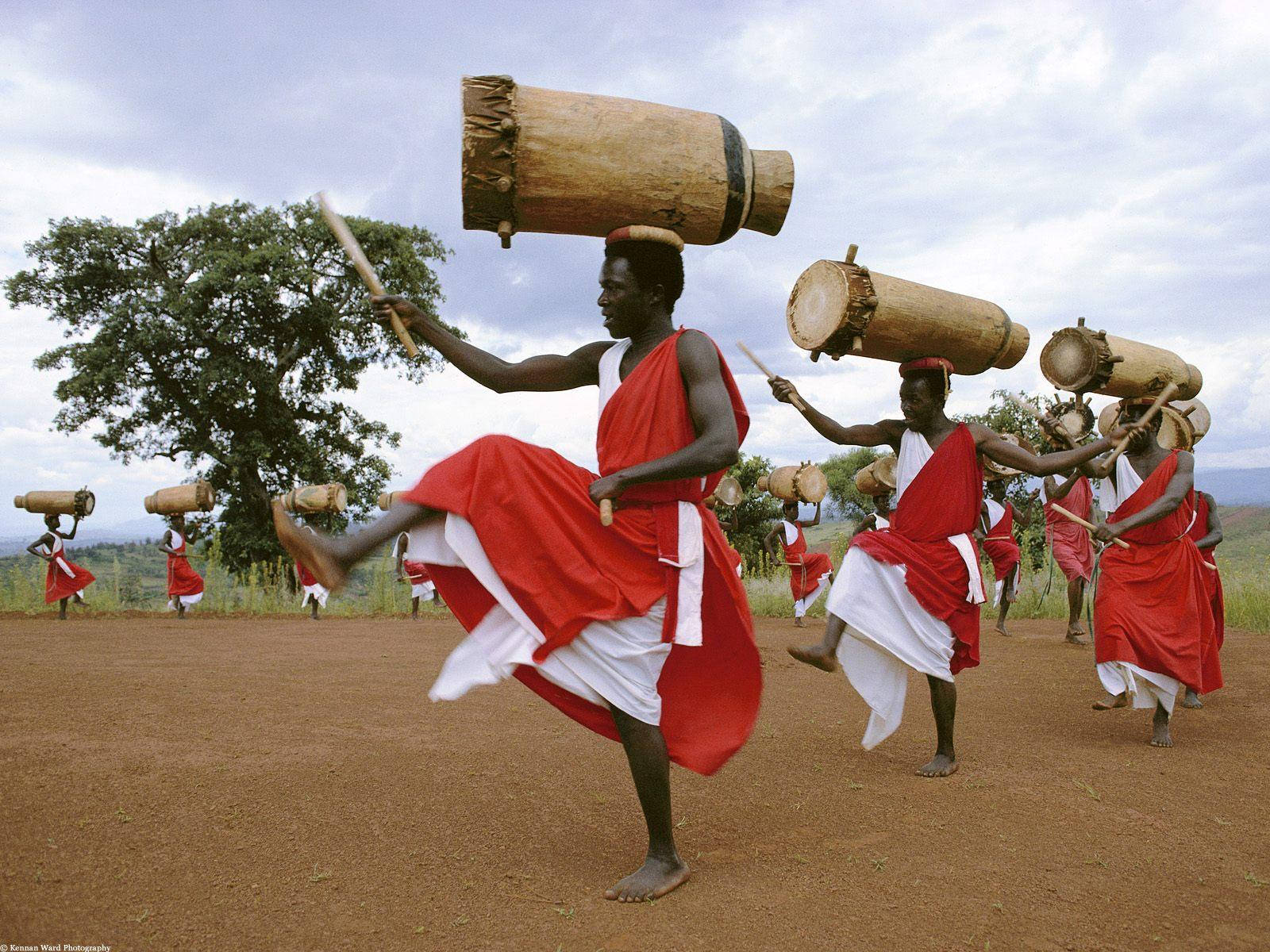 Africa 桌布 壁紙 — 身穿紅色和白色長袍的舞者在多雲的天空下，在泥地上表演動態的傳統舞蹈，同時頭頂平衡著大鼓。