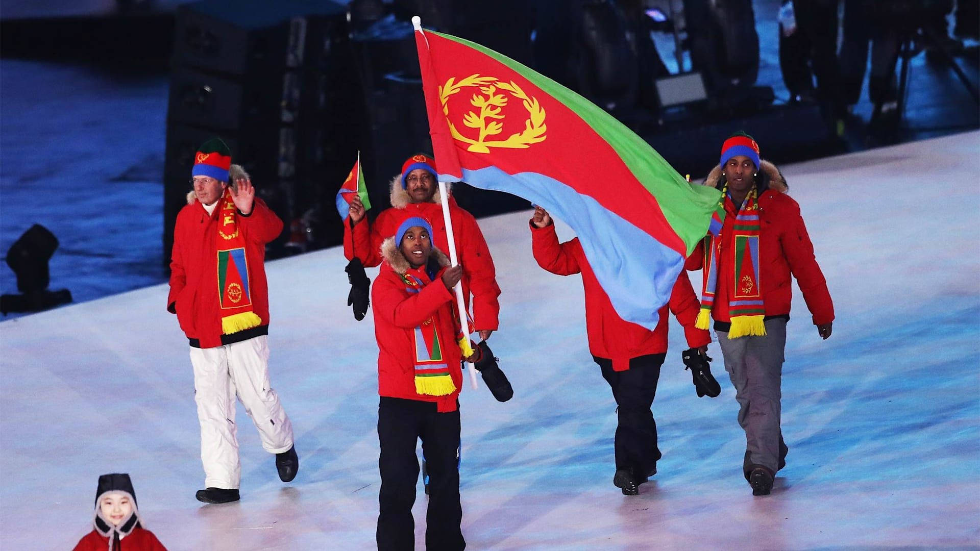 Eritrea 桌布 壁紙 — 身穿紅色外套的運動員自豪地高舉充滿活力的綠色、紅色和藍色厄立特里亞國旗，在冬季運動開幕式上行進。