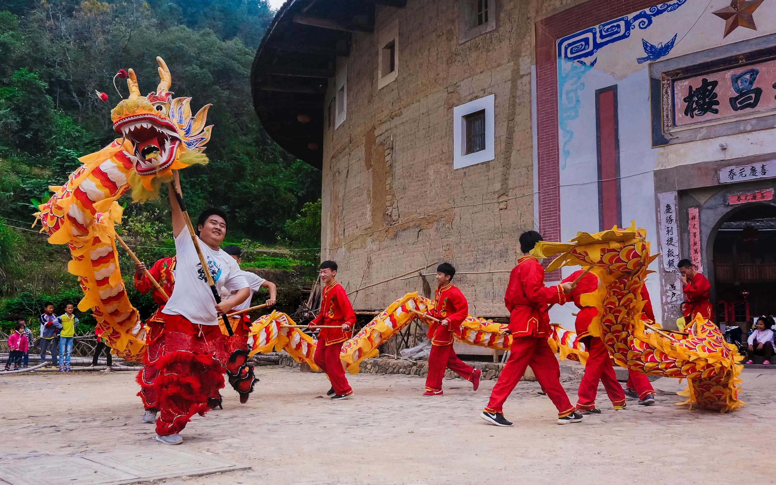 龍 桌布 壁紙 — 身穿紅色服裝的表演者在傳統中式土樓前舞動一條充滿活力的黃龍。