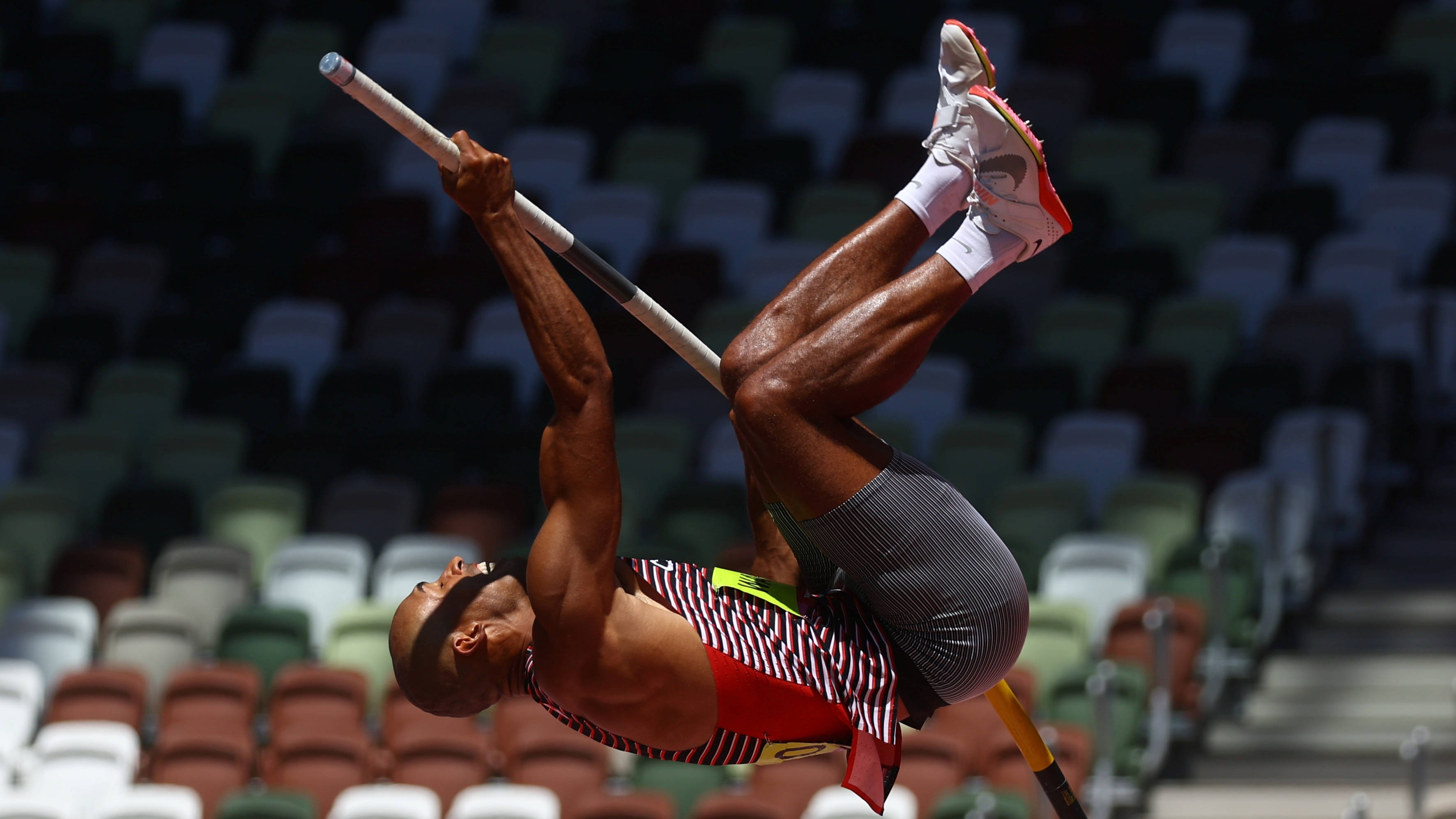 Athletic 桌布 壁紙 — 身穿紅色條紋制服的運動員在空蕩蕩的體育場座位背景下，在空中執行動態撐竿跳。