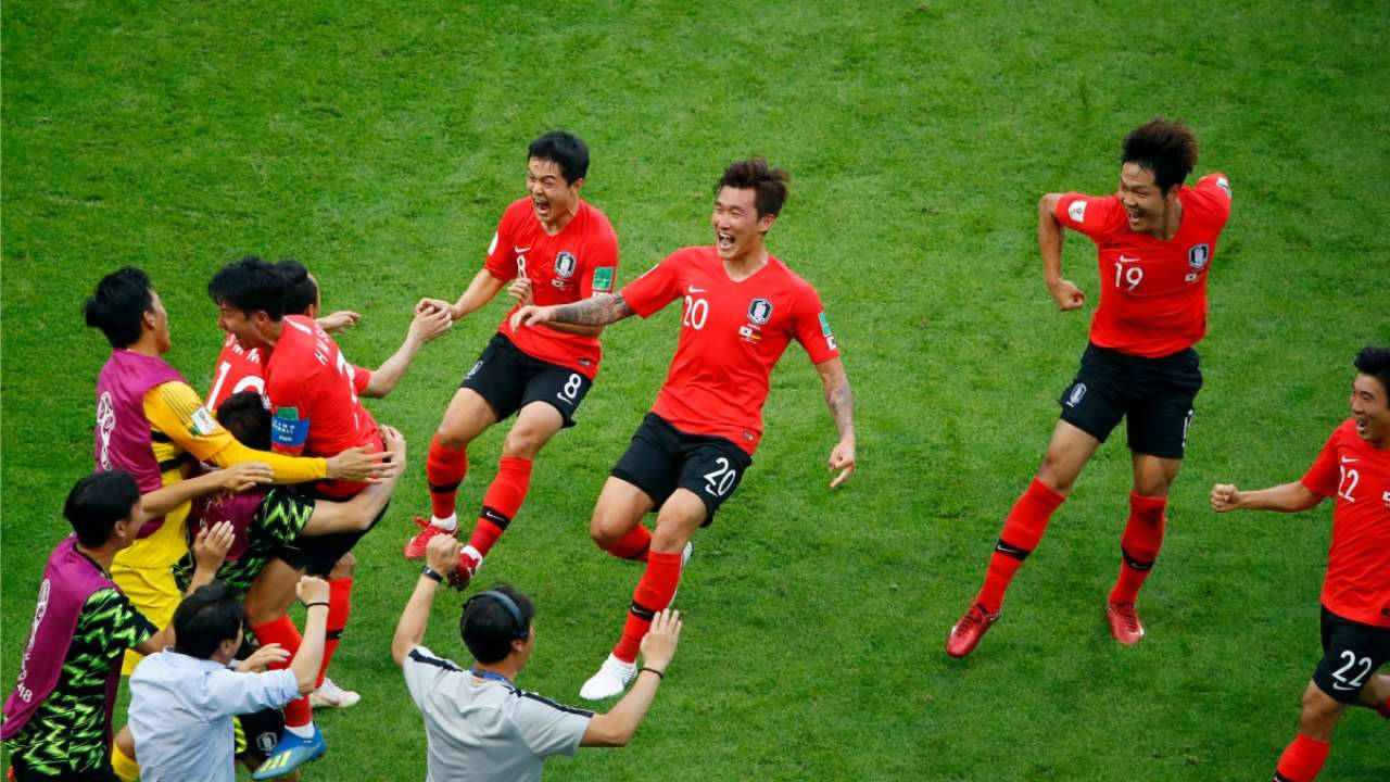 Football Players 桌布 壁紙 — 身穿紅色球衣的南韓足球員在充滿活力的綠色球場上狂喜慶祝進球。