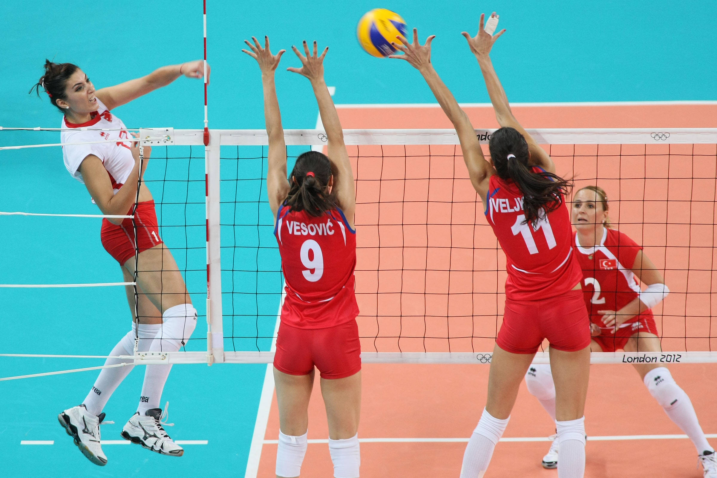 Indoor Volleyball 桌布 壁紙 — 身穿紅色球衣的女排球員在激烈的奧運比賽中跳起並攔截網前的黃色球。