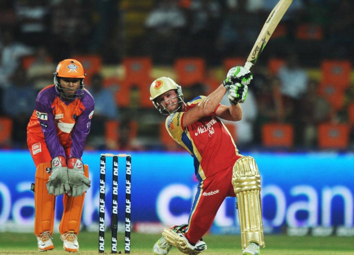 Ab De Villiers 桌布 壁紙 — 身穿紅色球衣的板球擊球手在球場燈光下強力揮棒，而身穿橙色裝備的守門員則蹲在門柱後面。