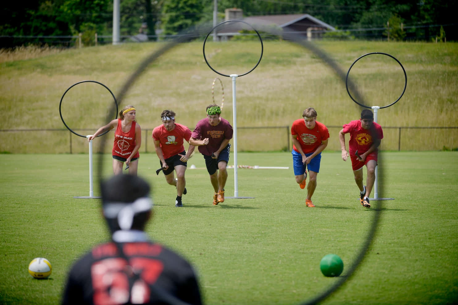 Quidditch 桌布 壁紙 — 身穿紅色球衣的球員在明亮的日光下，於戶外魁地奇比賽中在草地上奔跑躲避圓環。
