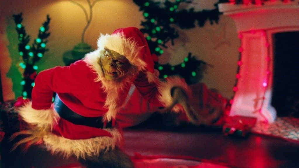 Grinch Zoom 桌布 壁紙 — 身穿紅色聖誕老人服裝的鬼靈精蹲在一個裝飾著發光綠色和紅色燈光的壁爐旁。
