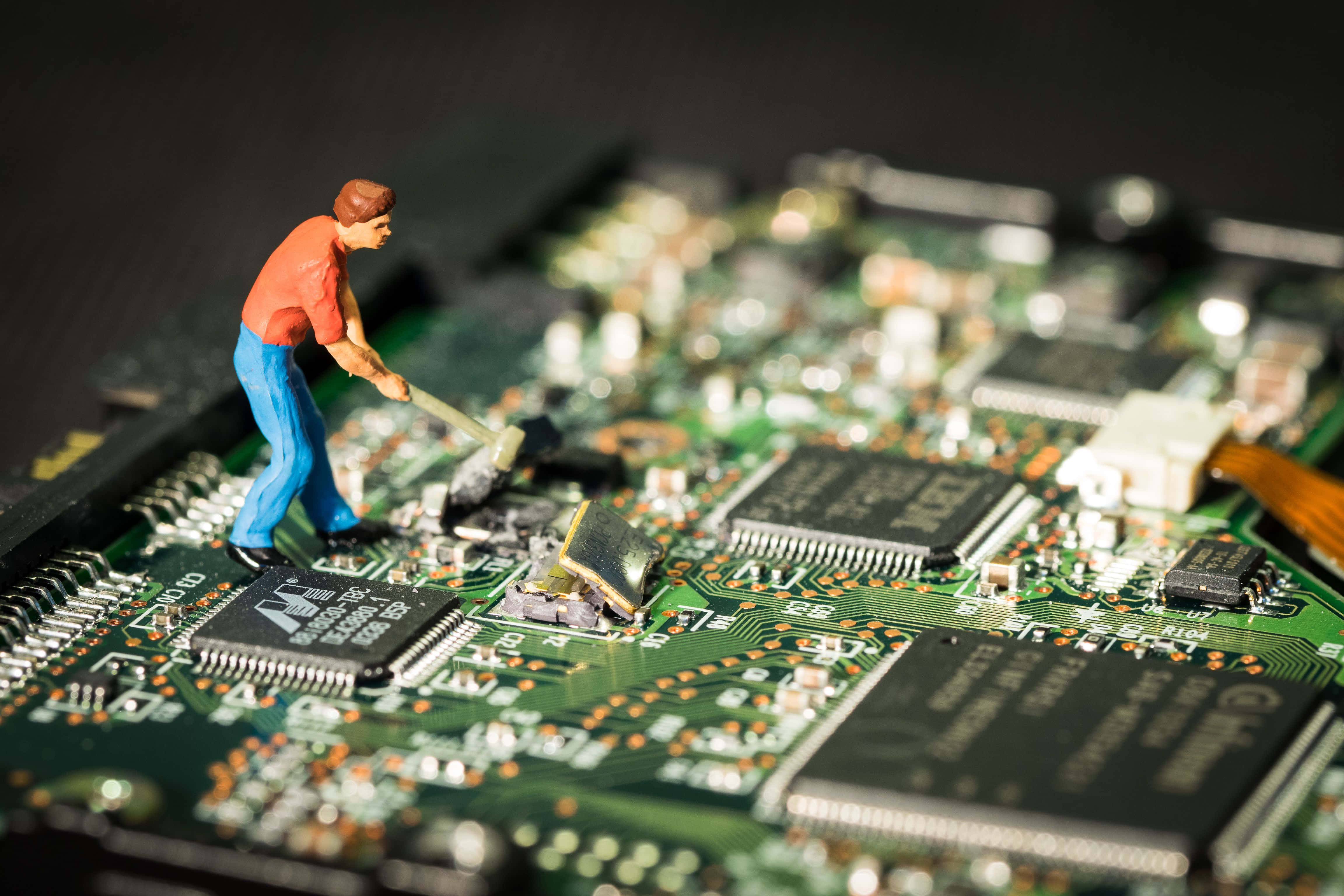Electronics 桌布 壁紙 — 身穿紅色襯衫和藍色褲子的小人偶用錘子敲擊詳細綠色電路板上的元件。
