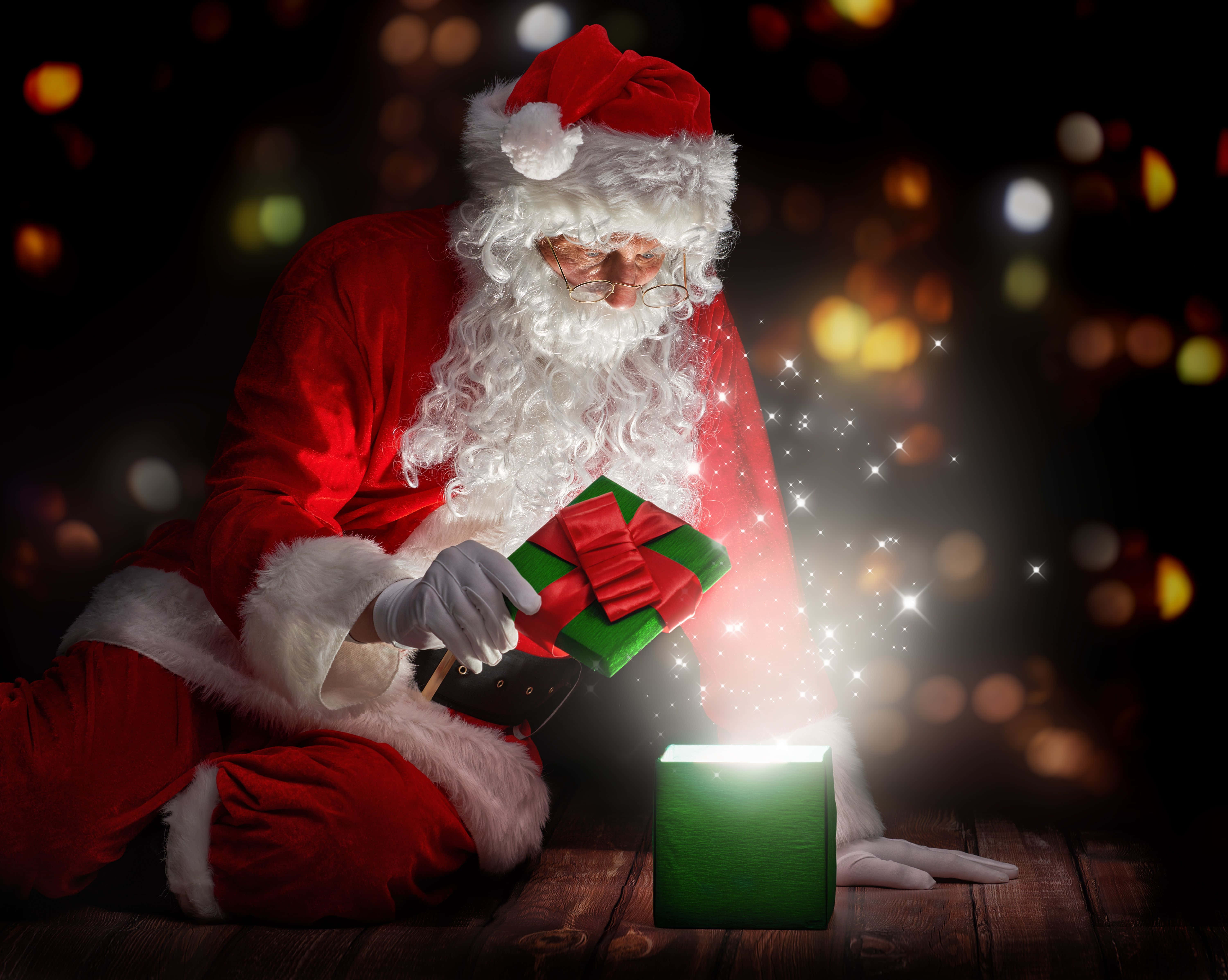 Santa Claus HD 桌布 壁紙 — 身穿紅色西裝的聖誕老人在木地板上，在散景燈光的背景下，打開一個帶有紅色蝴蝶結的發光綠色禮物盒。