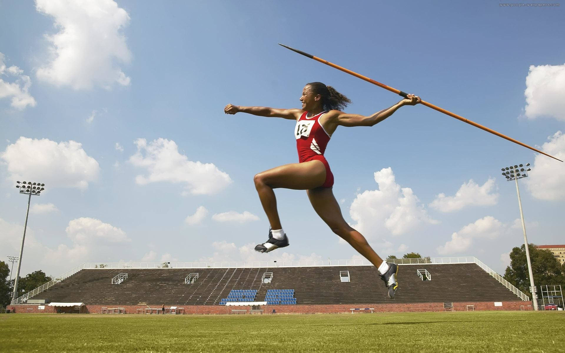 Athletic 桌布 壁紙 — 身穿紅色運動服的運動員在半空中擲標槍，背景是體育場上方明亮的藍天和蓬鬆的雲朵。