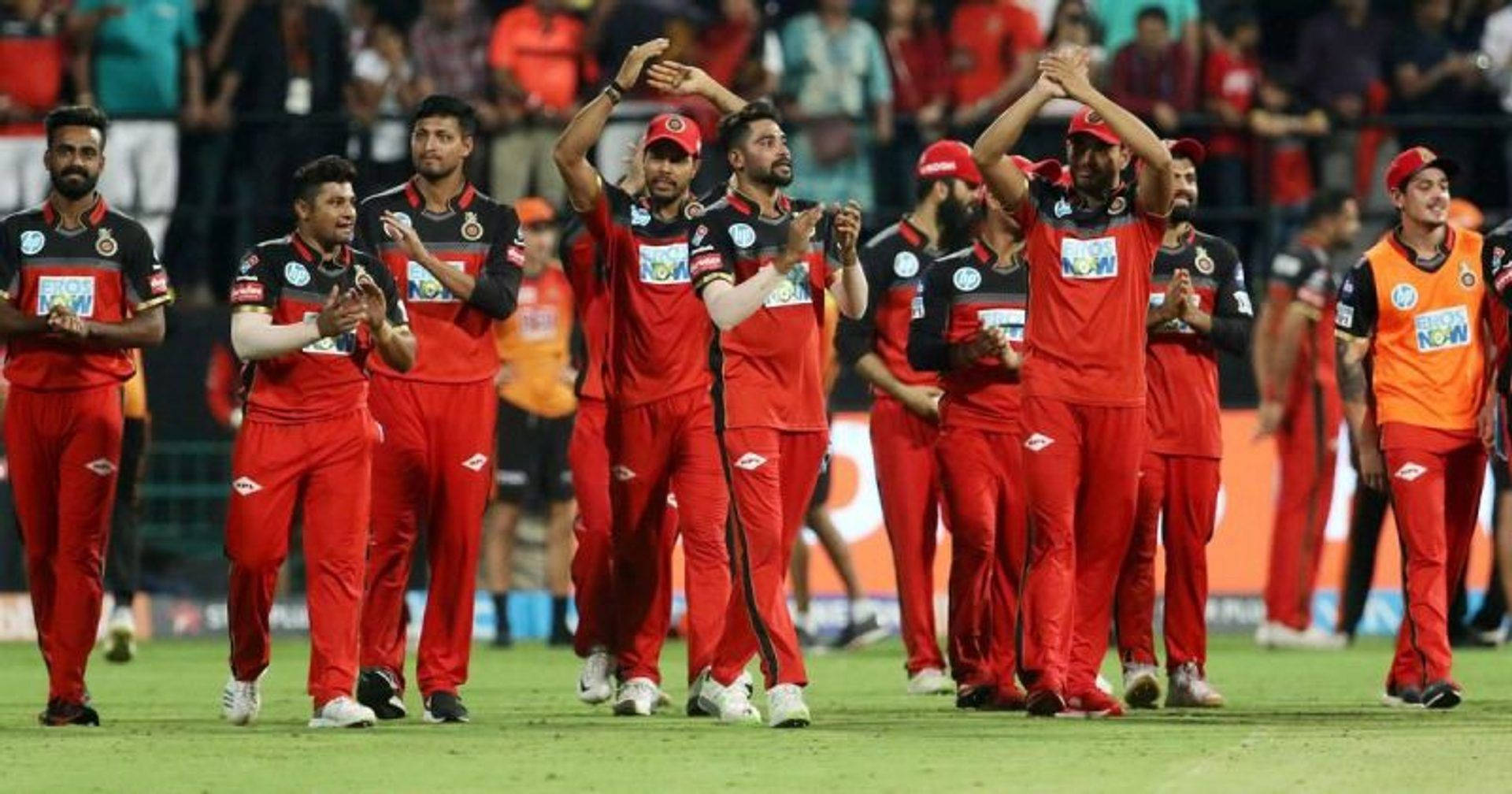 royal challengers bangalore 桌布 壁紙 — 身穿紅色隊服的板球隊在夜間體育場燈光下的綠色球場上為球迷鼓掌。