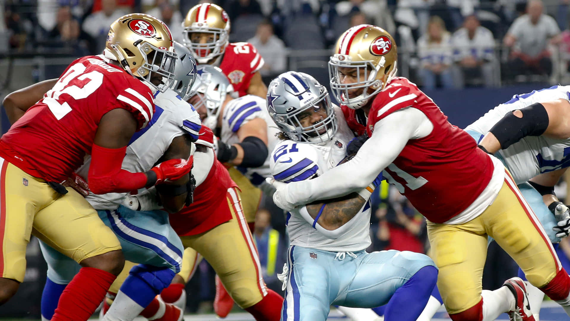 San Francisco 49ers 桌布 壁紙 — 身穿紅金色和淺藍色球衣的足球運動員在夜間體育場比賽中激烈碰撞。