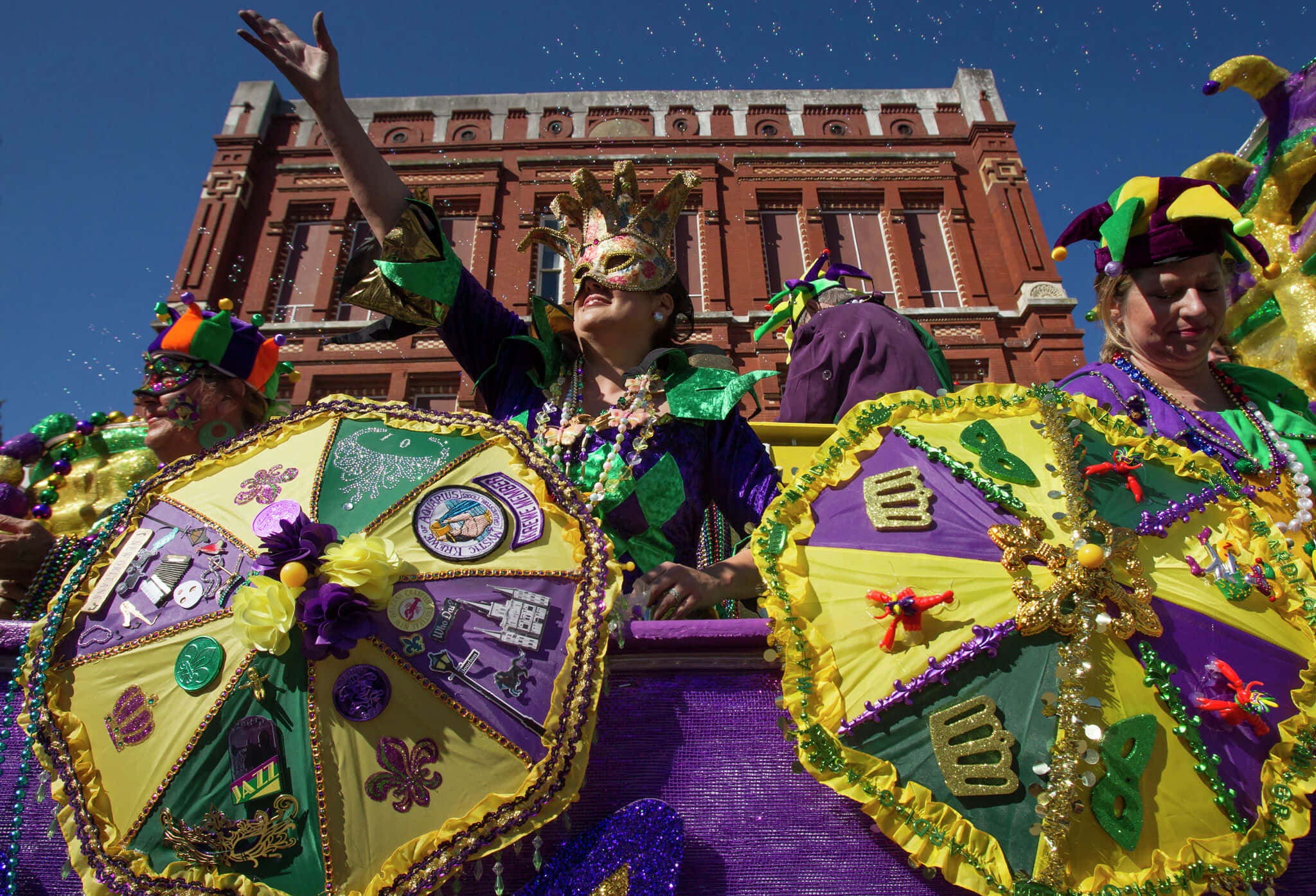 Mardi Gras 桌布 壁紙 — 身穿紫色、綠色和金色服裝的狂歡節狂歡者在晴朗的藍天下從裝飾花車上拋出珠子。
