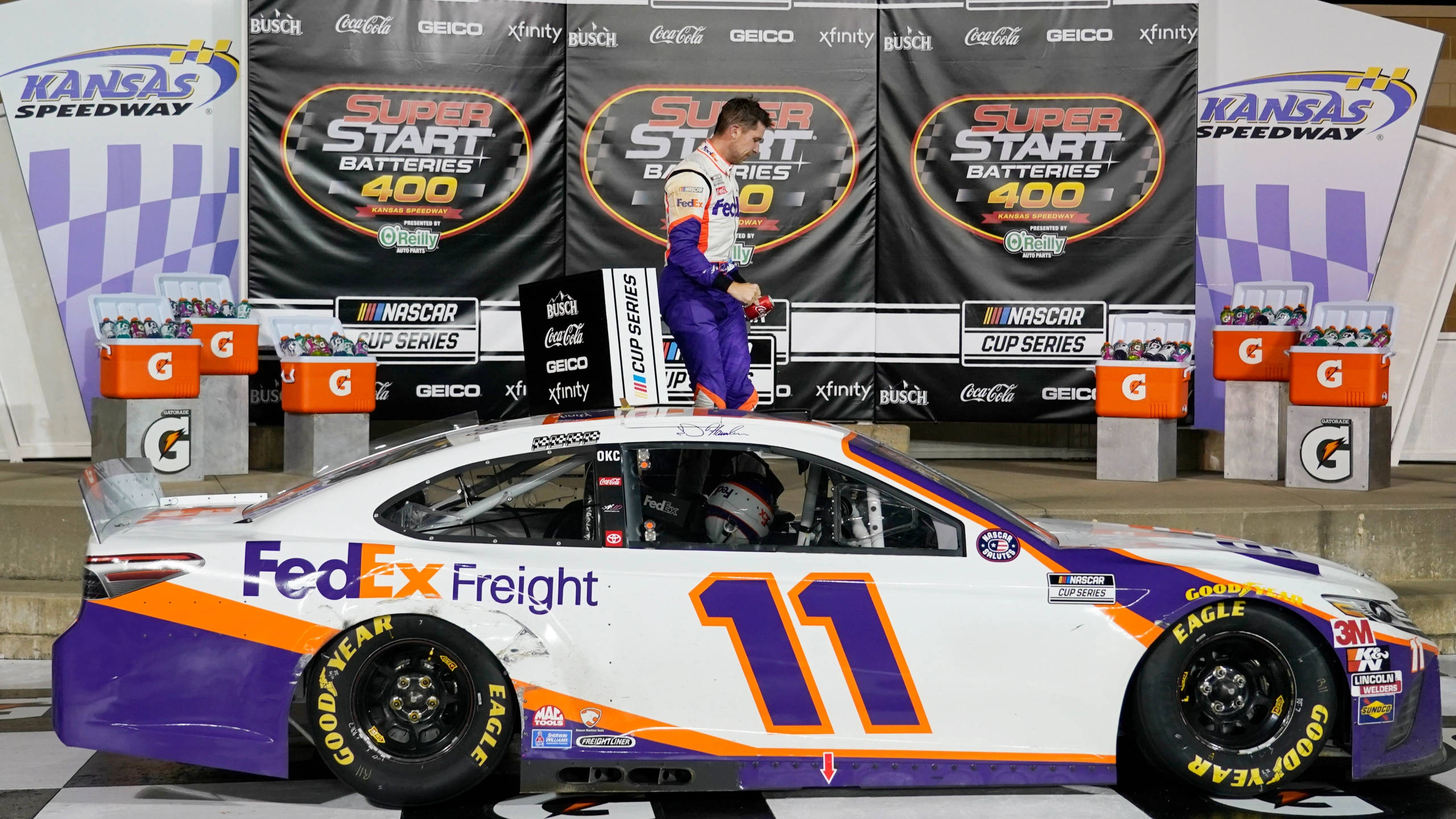Denny Hamlin 桌布 壁紙 — 身穿紫色和白色賽車服的NASCAR車手，站在堪薩斯賽車場頒獎台上他的FedEx Freight賽車頂部。