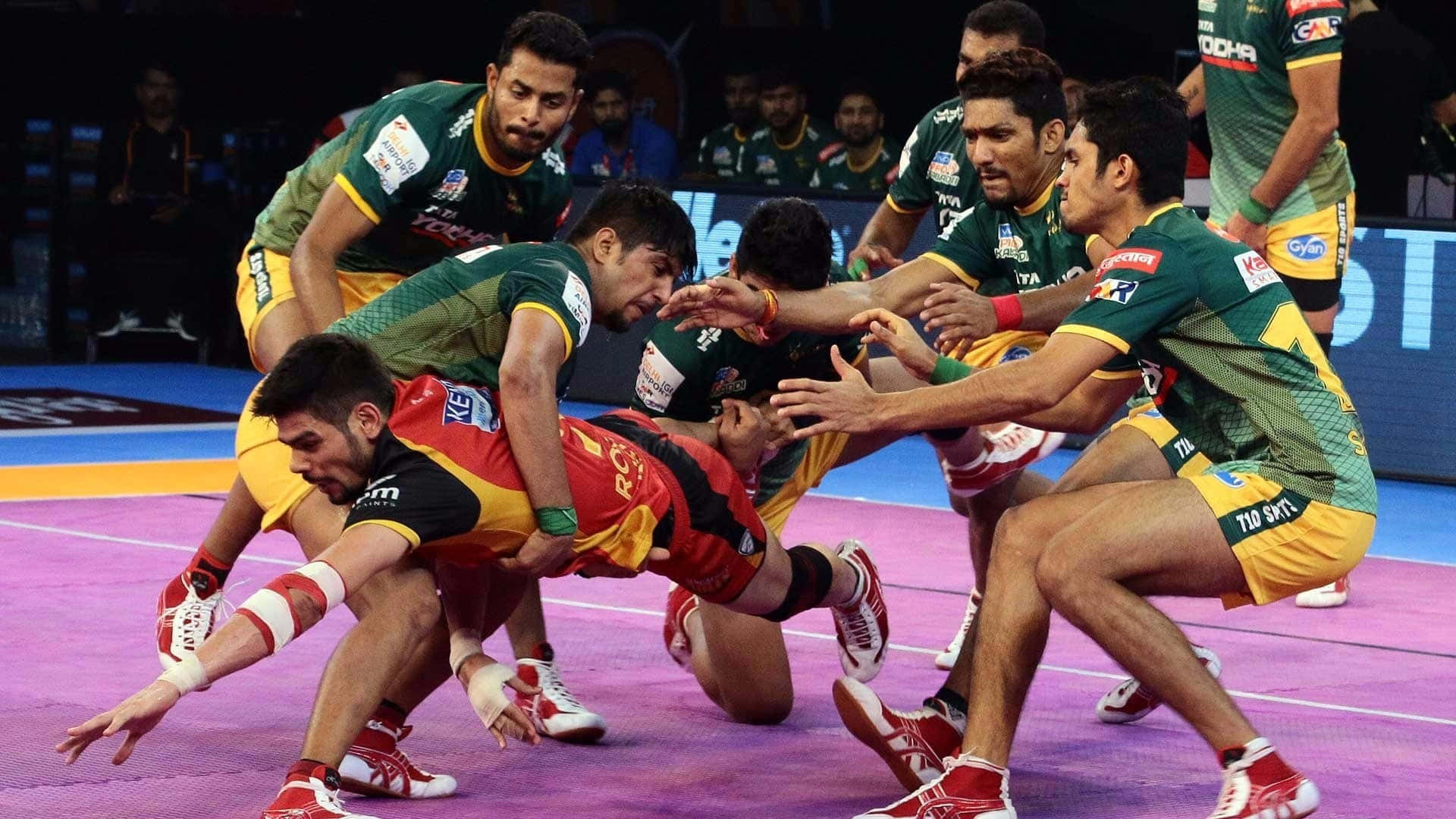 Kabaddi 桌布 壁紙 — 身穿綠色、黃色和紅色制服的激烈卡巴迪選手，在明亮的場館燈光下，在充滿活力的紫色場地上激烈搏鬥。