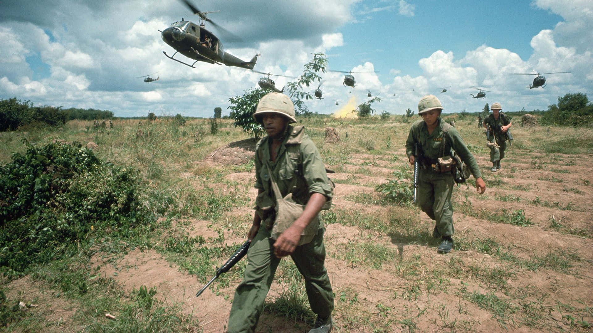 越南 桌布 壁紙 — 身穿綠色軍服的士兵在塵土飛揚的田野上行進，直升機在多雲的天空下從頭頂飛過。