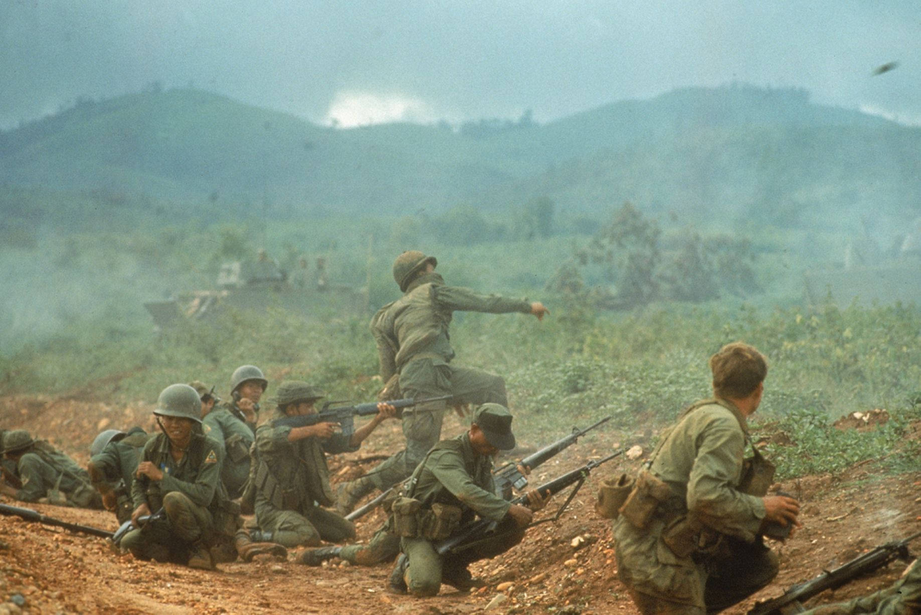 Hanoi 桌布 壁紙 — 身穿綠色軍裝的士兵蹲伏並穿過煙霧瀰漫的叢林戰場，遠處有山丘和煙霧。