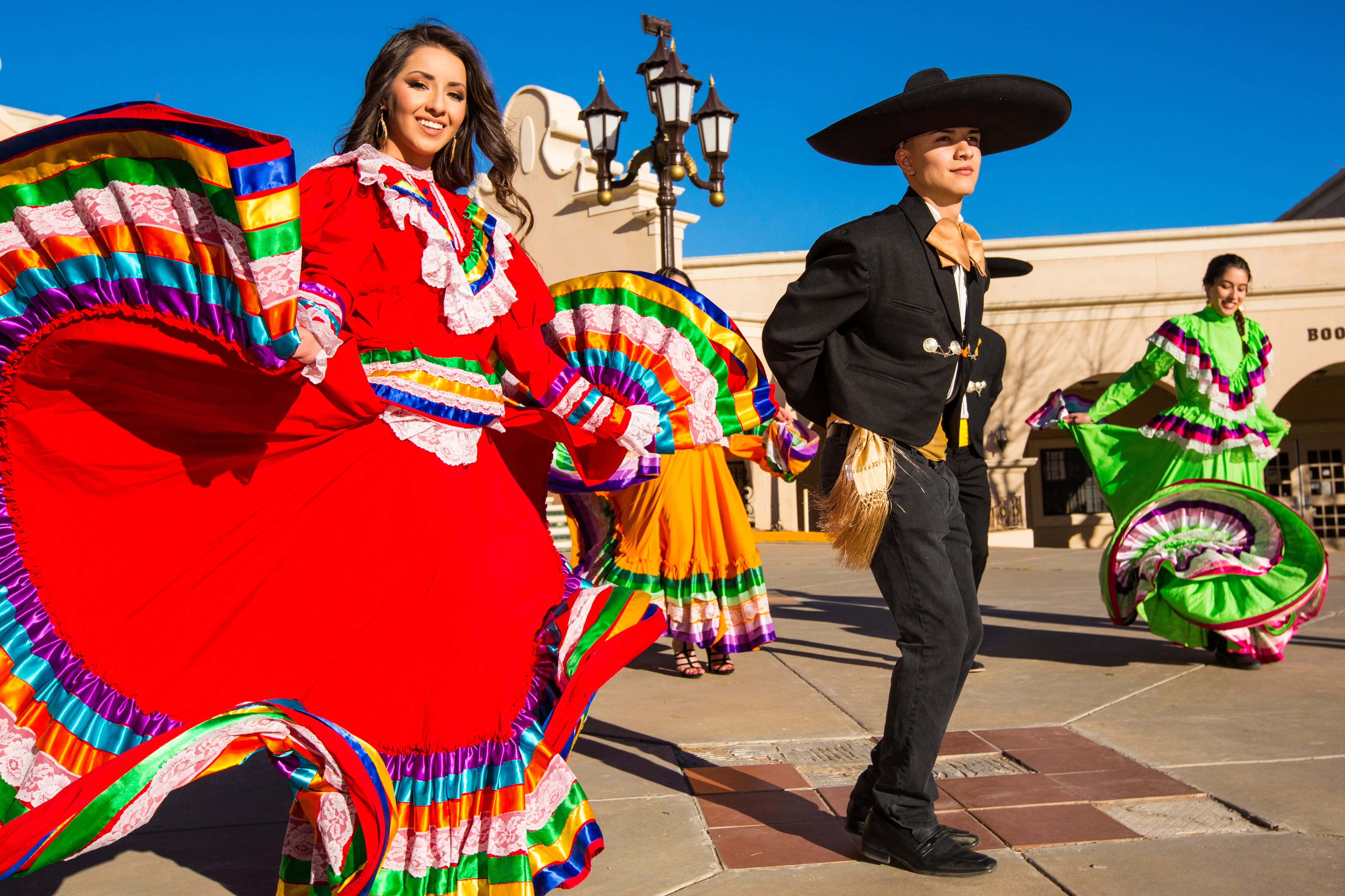 Mexican 桌布 壁紙 — 身穿色彩繽紛傳統服飾的活力舞者在陽光明媚的廣場上表演生動的民間舞蹈，廣場上有華麗的路燈和石鋪路面。
