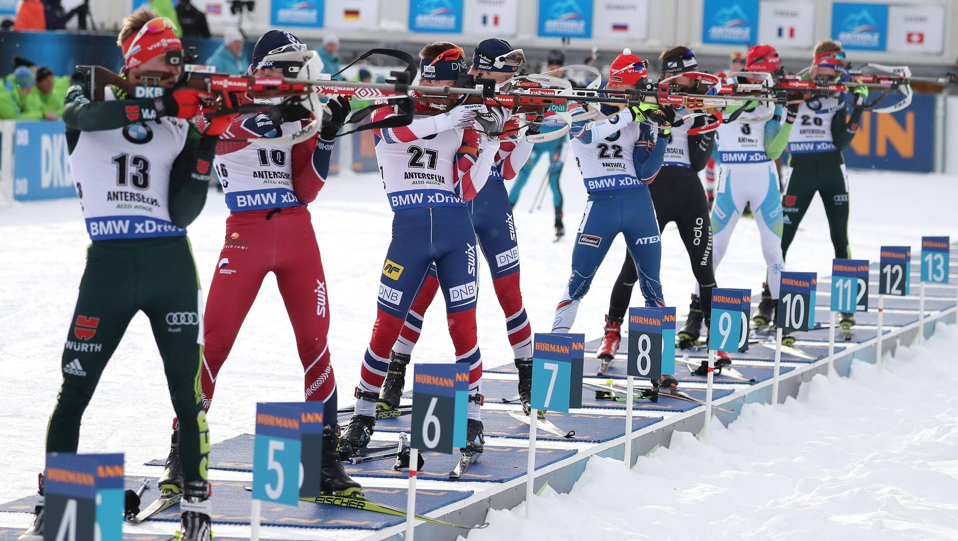 Winter Olympics 桌布 壁紙 — 身穿色彩繽紛制服的冬季兩項運動員在雪地射擊場瞄準步槍，進行一場競爭激烈的冬季賽事。