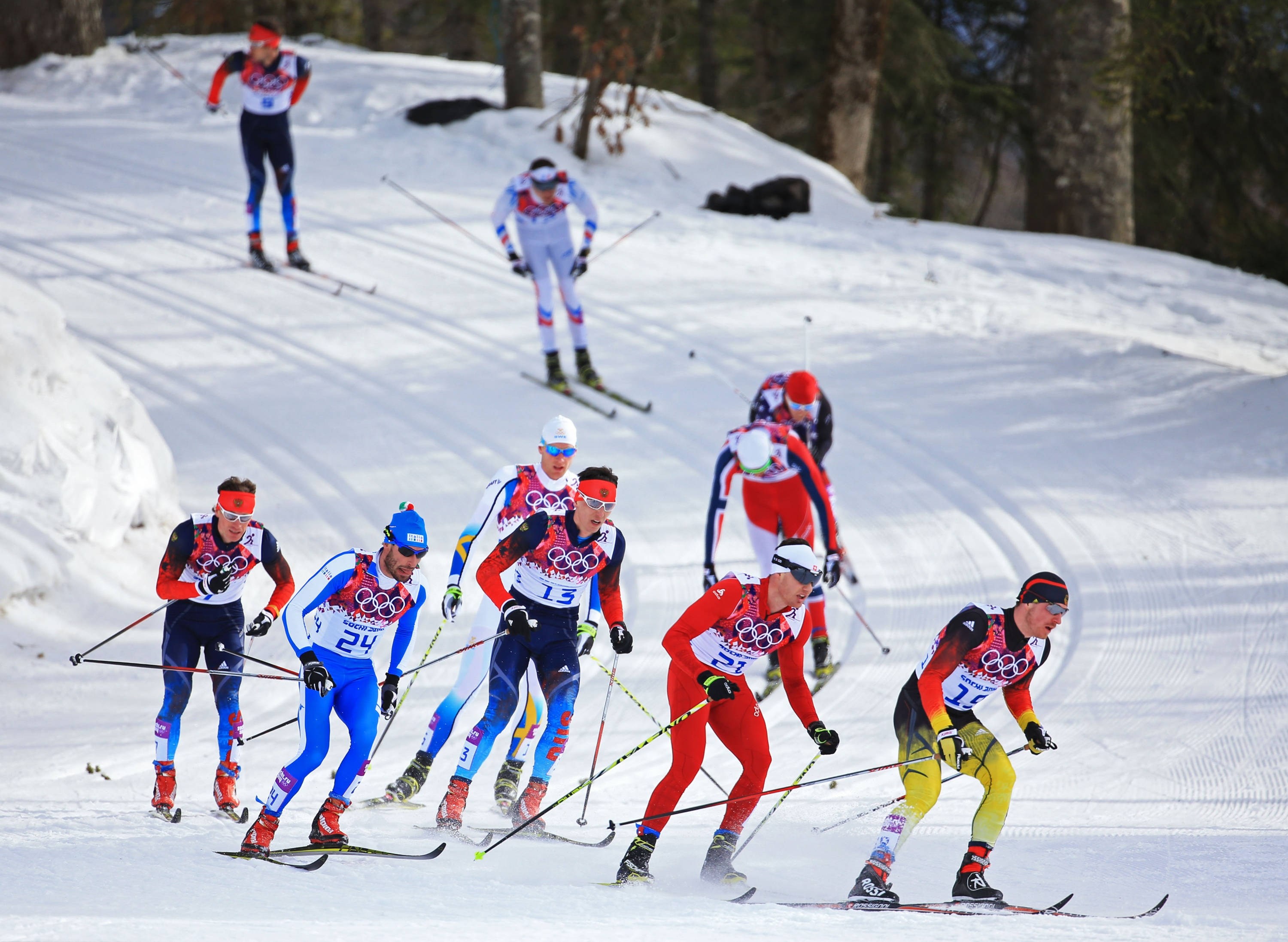 Winter Olympics 桌布 壁紙 — 身穿色彩繽紛奧運制服的越野滑雪選手在白雪覆蓋的森林小徑上競速。