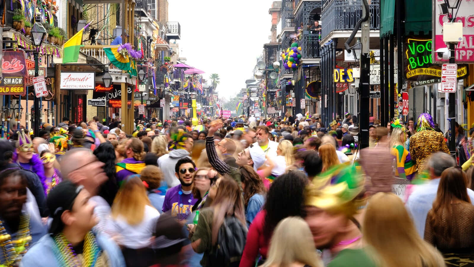 Mardi Gras 桌布 壁紙 — 身穿色彩繽紛狂歡節服飾和珠串的狂歡人群，擠滿了一條充滿節日氣氛的街道，兩旁是裝飾華麗的建築物和懸掛的旗幟。
