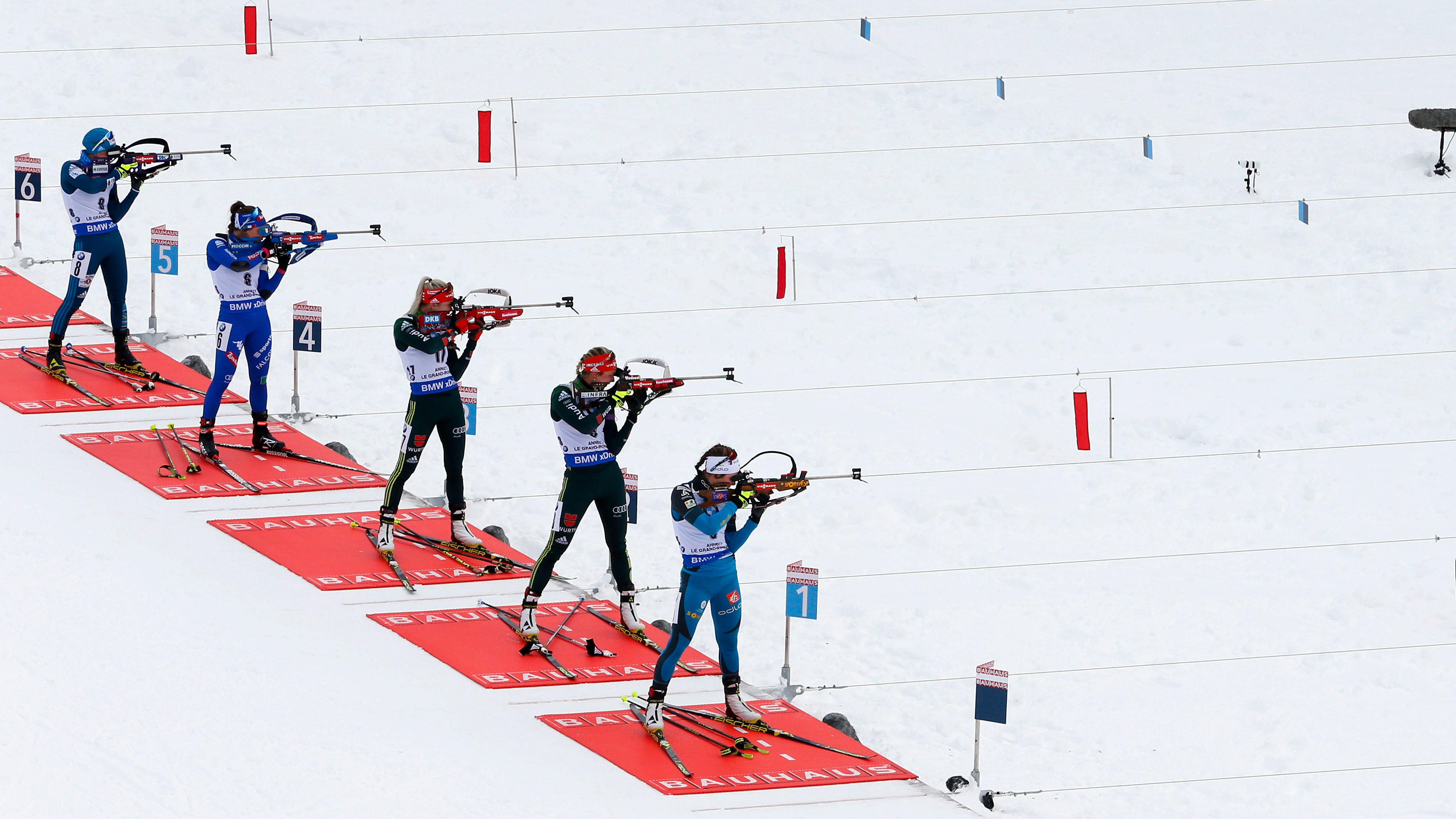 運動 桌布 壁紙 — 身穿色彩繽紛裝備的冬季兩項運動員在雪地上，從紅色射擊墊上瞄準步槍，賽道標有編號靶標。