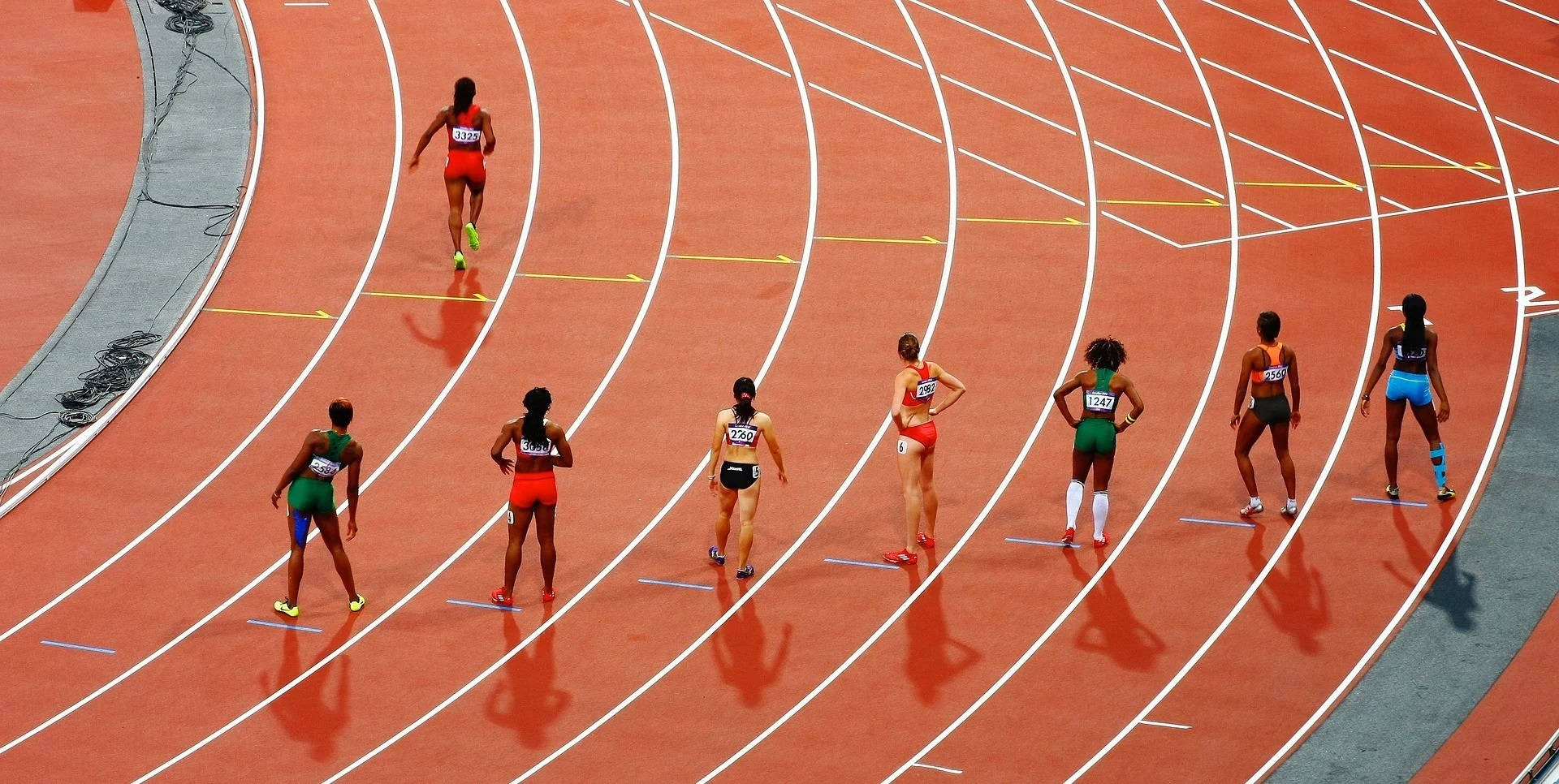 Olympics 桌布 壁紙 — 身穿色彩繽紛運動服的運動員，在明亮的日光下，站在有白色跑道的彎曲紅色跑道上蓄勢待發。