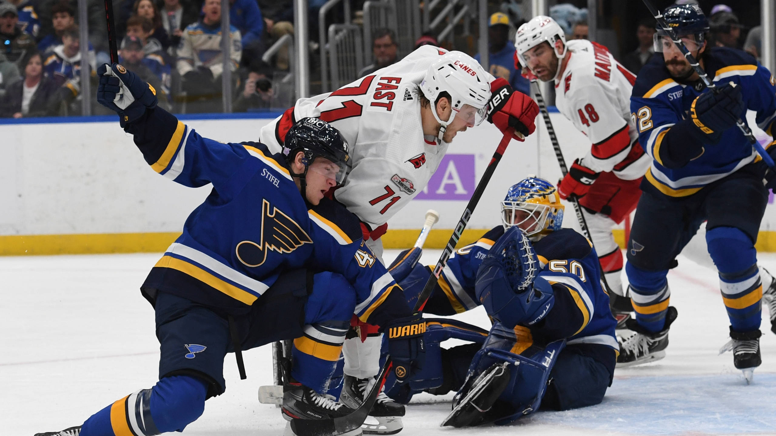 St Louis Blues 桌布 壁紙 — 身穿藍白球衣的冰球運動員在快速比賽中圍繞球門激烈爭奪，背景有觀眾。