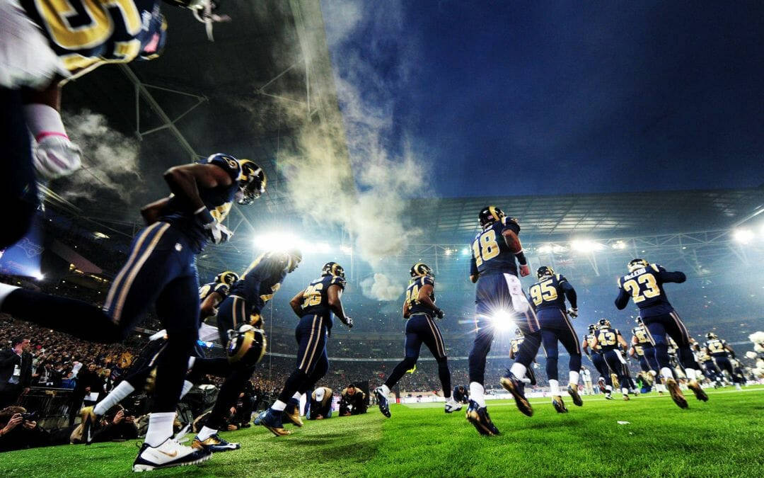 NFL 桌布 壁紙 — 身穿藍色和金色球衣的足球運動員在體育場燈光下衝入球場，身後煙霧翻騰。