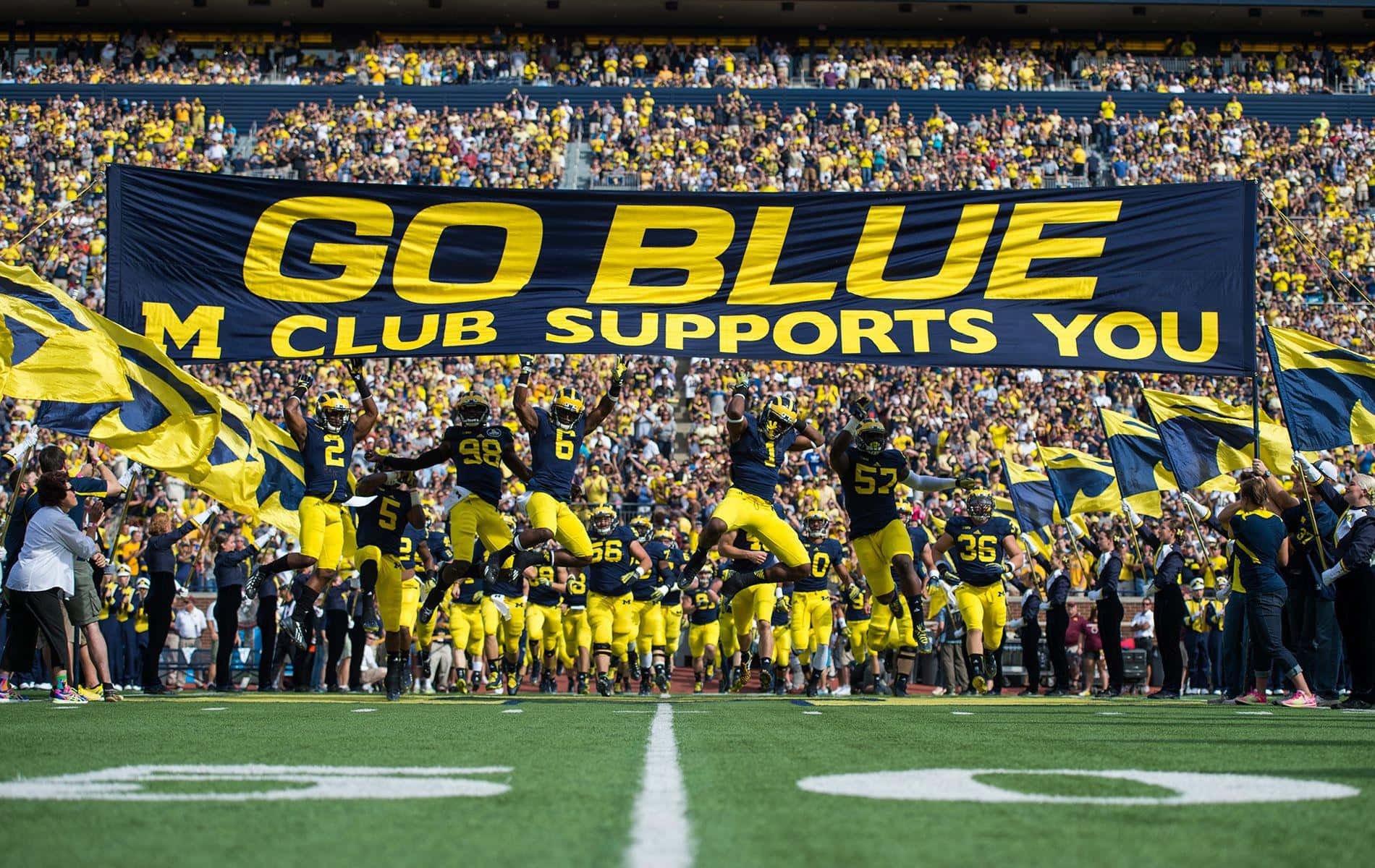 Michigan Football 桌布 壁紙 — 身穿藍色和黃色球衣的足球員跳入球場，球迷揮舞旗幟，一個大橫幅寫著「GO BLUE M CLUB SUPPORTS YOU」。