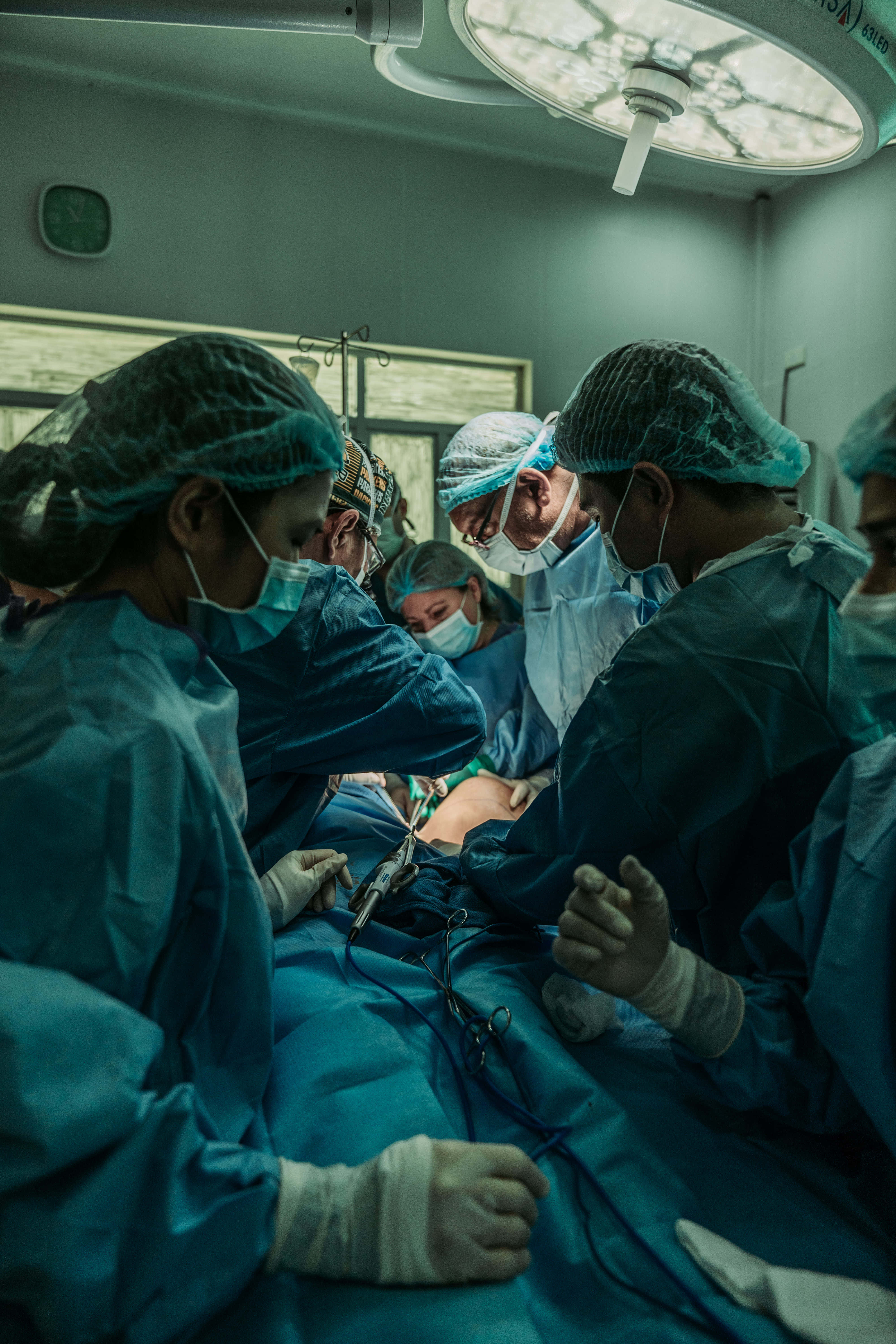 Hospital 桌布 壁紙 — 身穿藍色手術服和口罩的外科醫生在明亮的手術燈光下專注於病人。