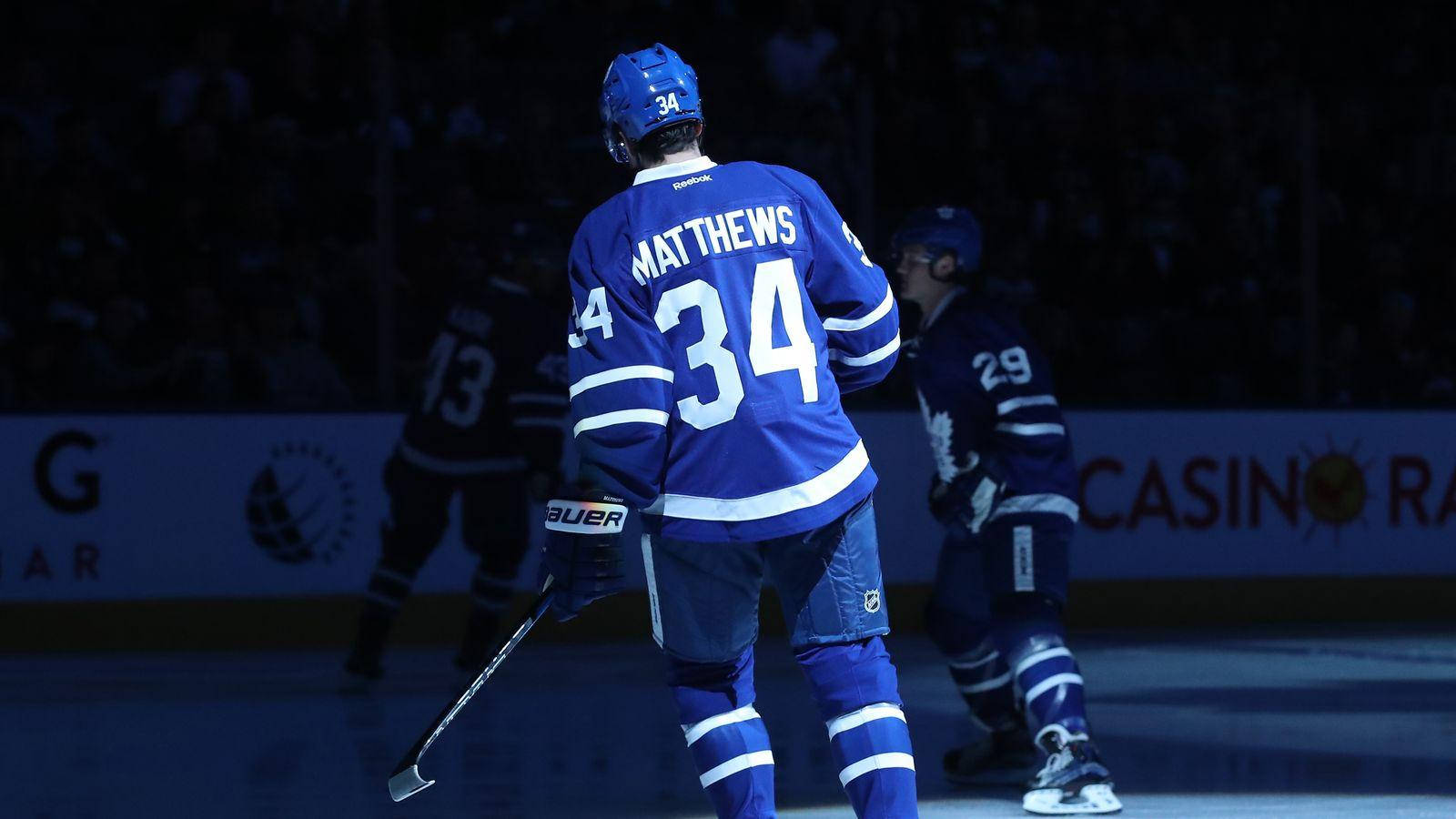 運動 桌布 壁紙 — 身穿藍色球衣的冰球運動員 Matthews 34號在比賽期間站在冰場上，場館燈光照射下。