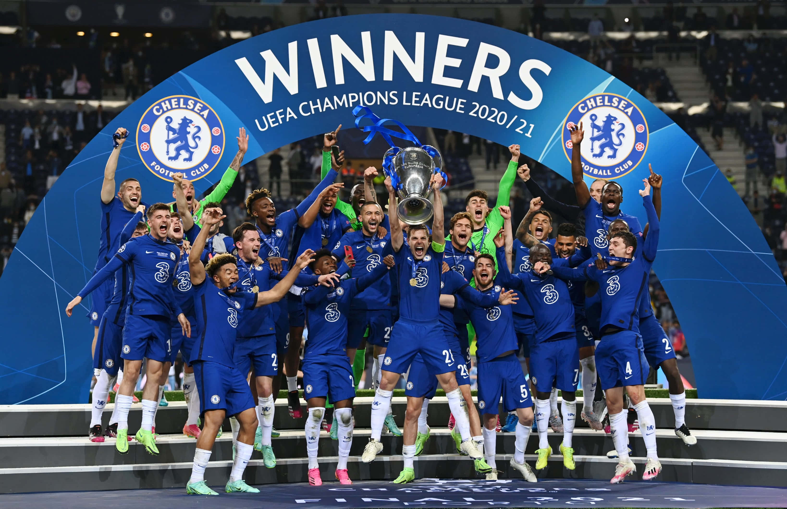 Champions League 桌布 壁紙 — 身穿藍色球衣的切爾西足球員在明亮的球場燈光下，在頒獎台上歡樂跳躍，高舉歐冠冠軍獎盃。