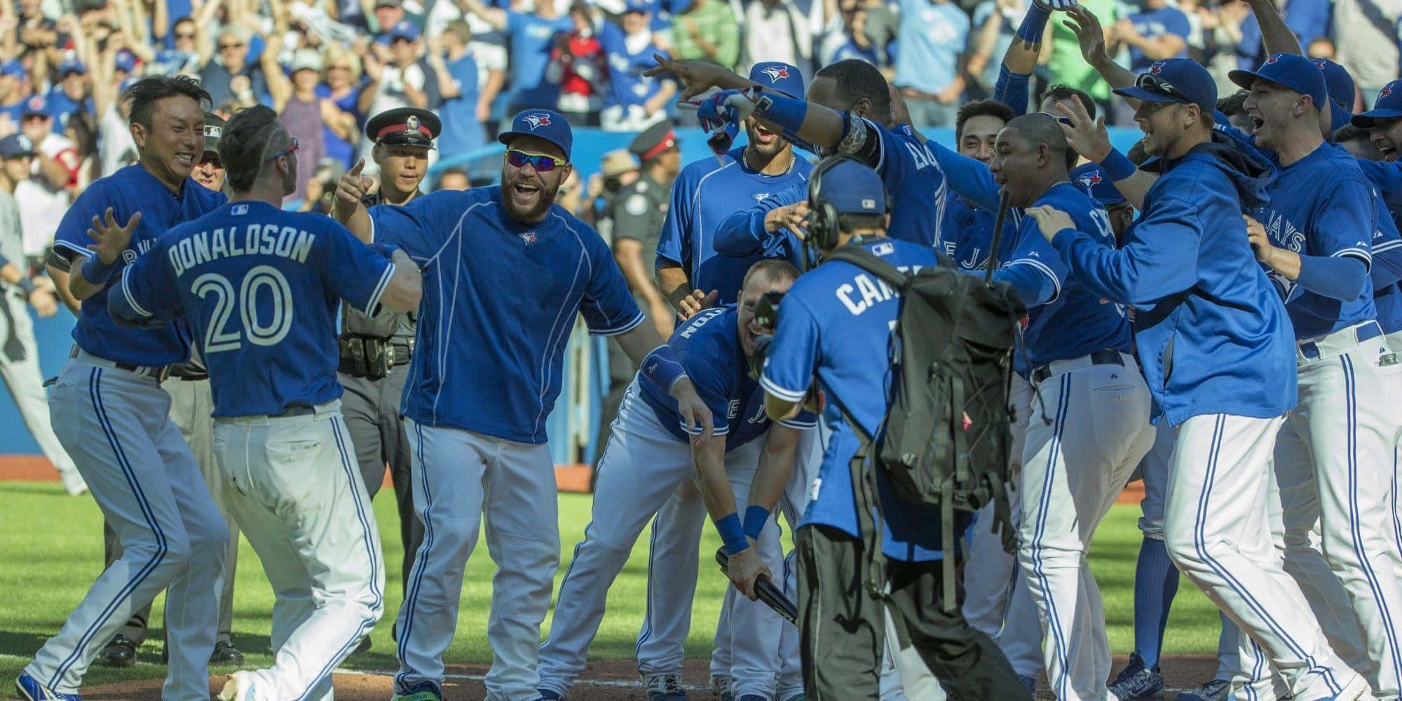 Blue Jays. 桌布 壁紙 — 身穿藍色球衣的棒球運動員在陽光明媚的球場上與背景中的球迷一起興高采烈地慶祝。