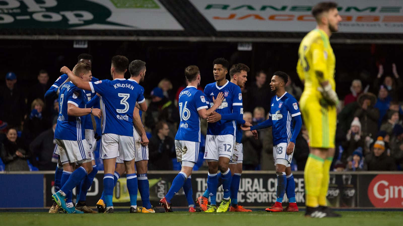 Ipswich Town 桌布 壁紙 — 身穿藍色球衣的足球運動員在燈光璀璨的球場上慶祝進球，附近有一位身穿黃色球衣的守門員。