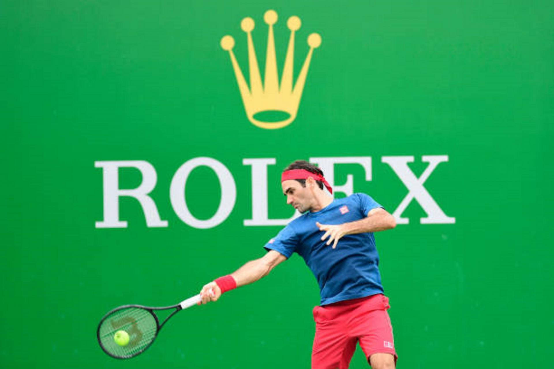 roger 桌布 壁紙 — 身穿藍色襯衫和紅色短褲的網球選手，在綠色勞力士牆前揮拍擊打黃色球。