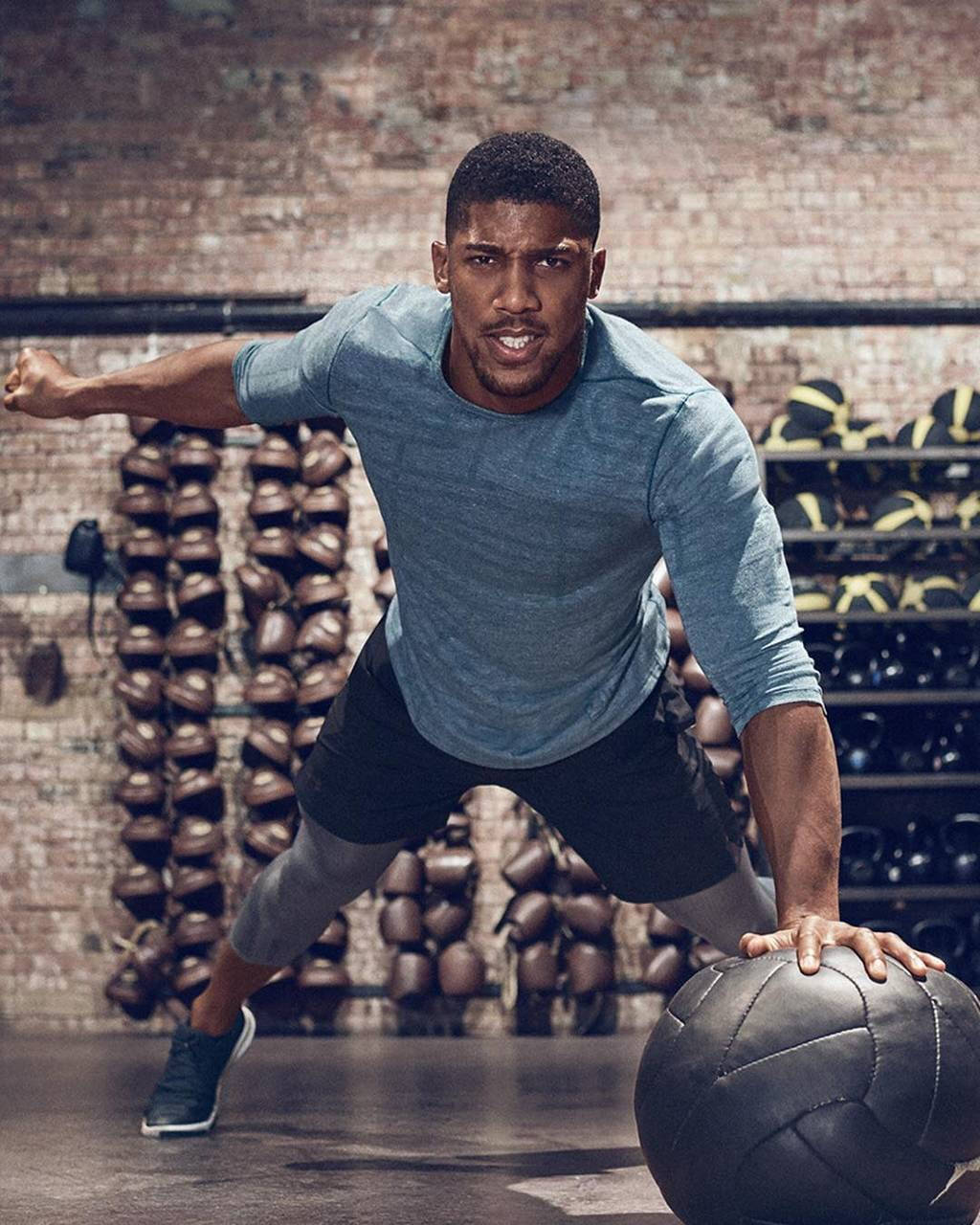 Anthony Joshua 桌布 壁紙 — 身穿藍色襯衫的運動型男子在粗糙的磚牆健身房中向前弓步，抓住一個黑色藥球。