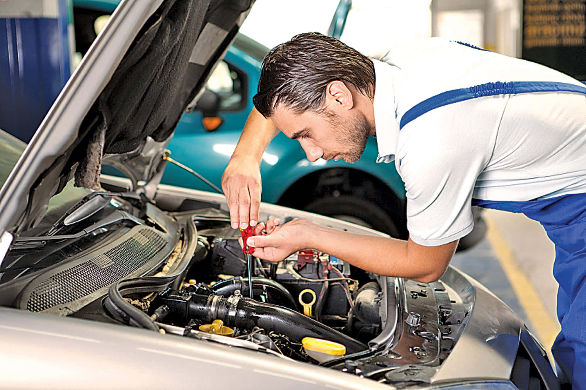 Car Repair 桌布 壁紙 — 身穿藍色連身工作服的機械師在車庫中，用紅色螺絲起子調整銀色汽車打開的引擎蓋下的引擎部件。