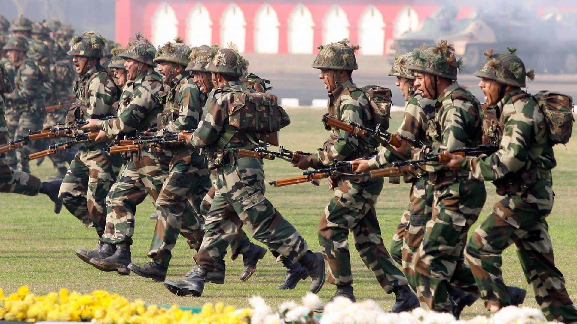 印度軍隊 桌布 壁紙 — 身穿迷彩服的士兵們手持步槍，整齊劃一地穿過草地，前景有黃色和白色的花朵。