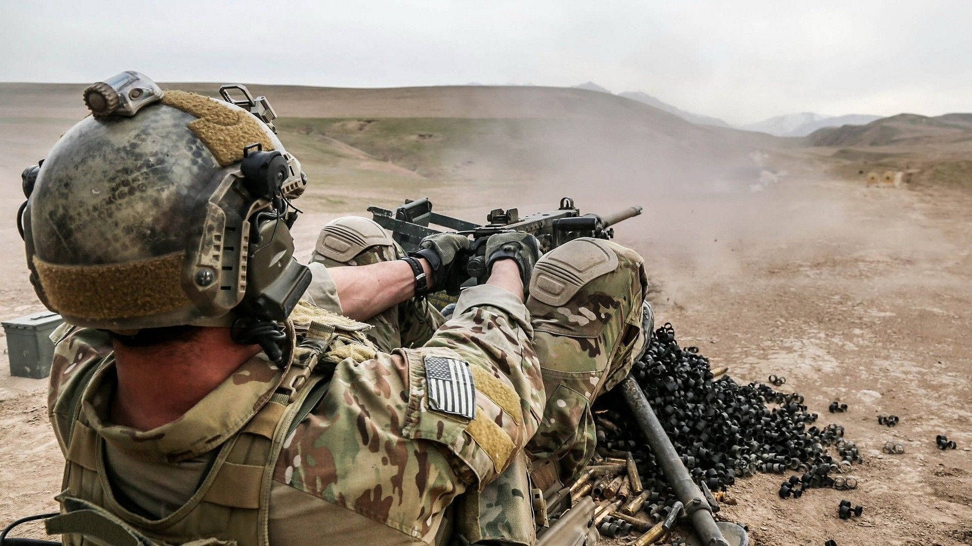 US 桌布 壁紙 — 身穿迷彩服的士兵在塵土飛揚的沙漠景觀中發射重型機槍，附近散落著用過的彈藥。
