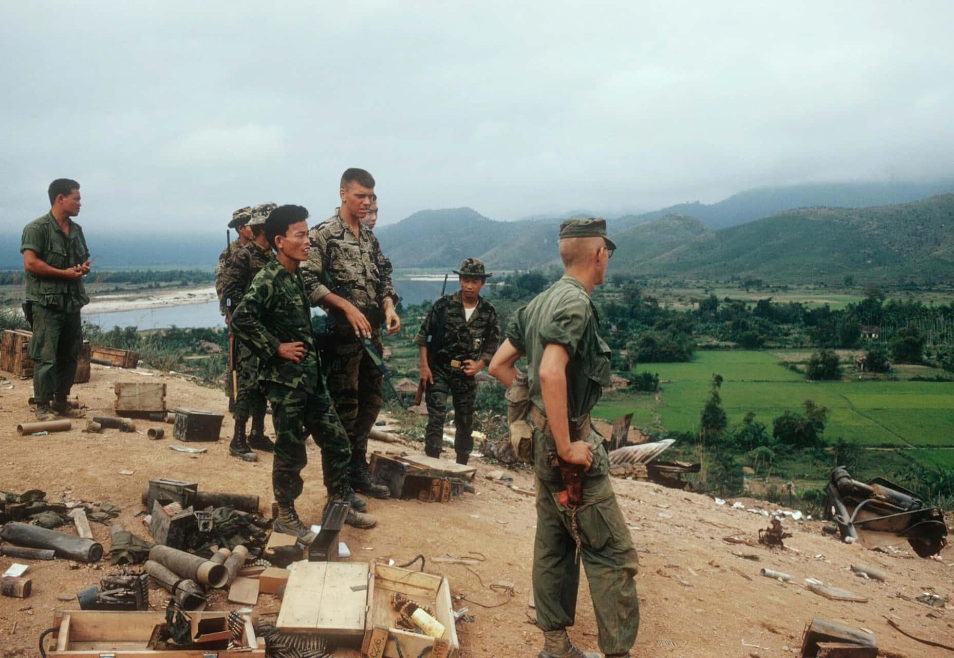 Vietnam War 桌布 壁紙 — 身穿迷彩服的士兵站在一個散落著彈藥箱和碎片的山丘上，俯瞰著一個有綠色田野和遠處山脈的山谷，天空多雲。