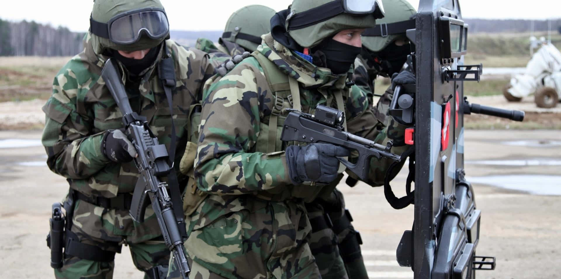 Special Forces 桌布 壁紙 — 身穿迷彩裝備和面具的士兵在開闊的田野中謹慎前進，手持步槍和盾牌。