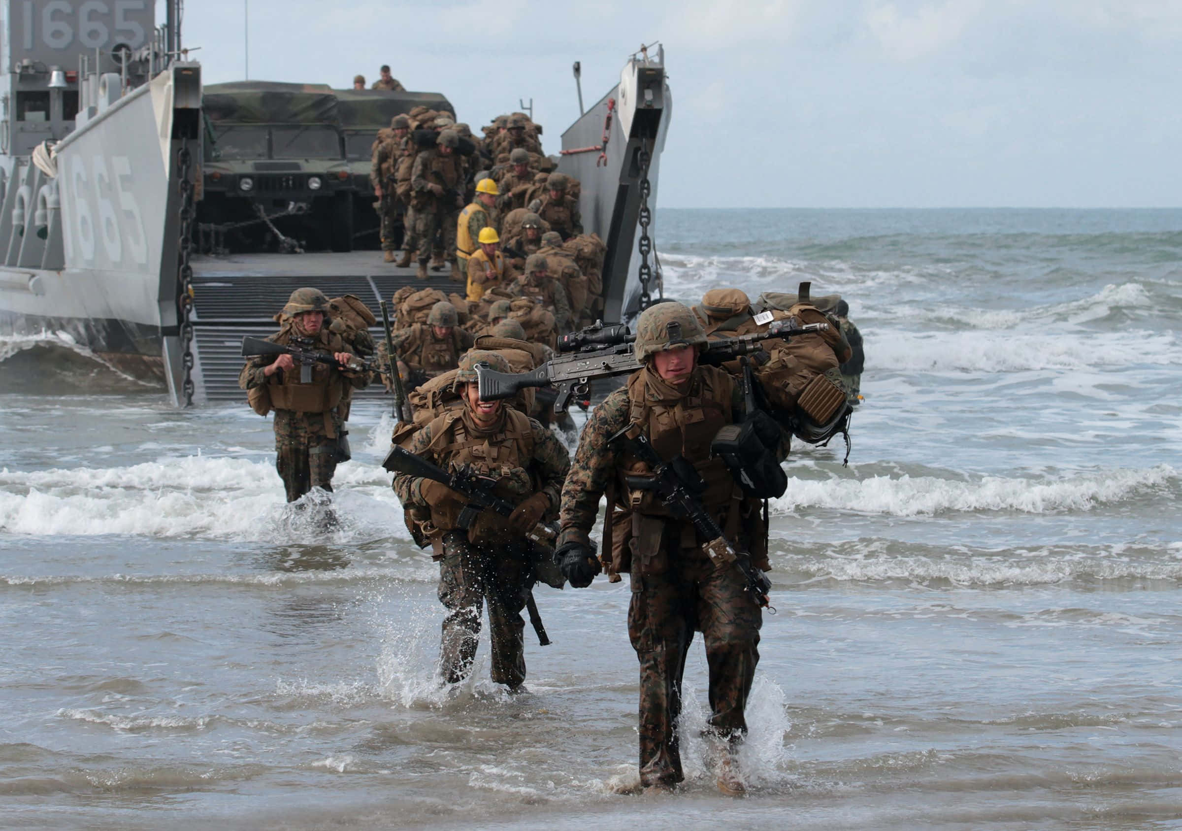 Marines 桌布 壁紙 — 身穿迷彩裝備的士兵在淺海水中涉水前進，攜帶重型裝備從軍用登陸艇下船。