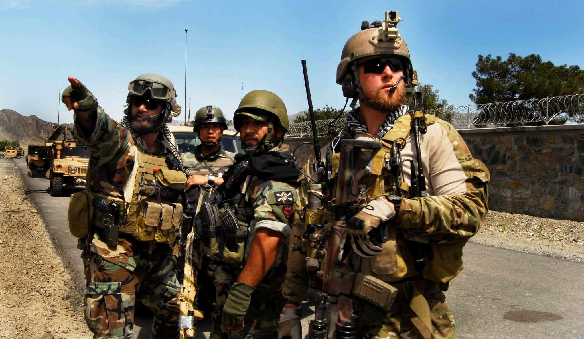 Special Forces 桌布 壁紙 — 身穿迷彩裝備的士兵在路邊警戒，旁邊有軍用車輛和鐵絲網圍欄，頭頂是明亮的藍天。