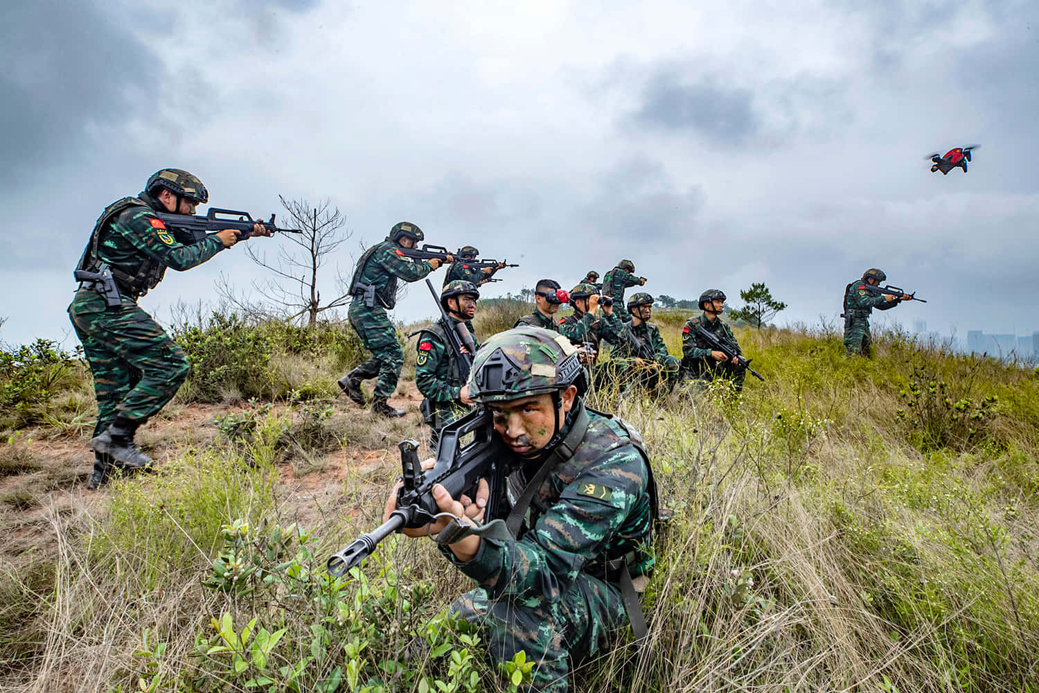 戰爭動圖 桌布 壁紙 — 身穿迷彩裝備的士兵在陰天天空下穿越草坡，一架無人機在頭頂飛過。