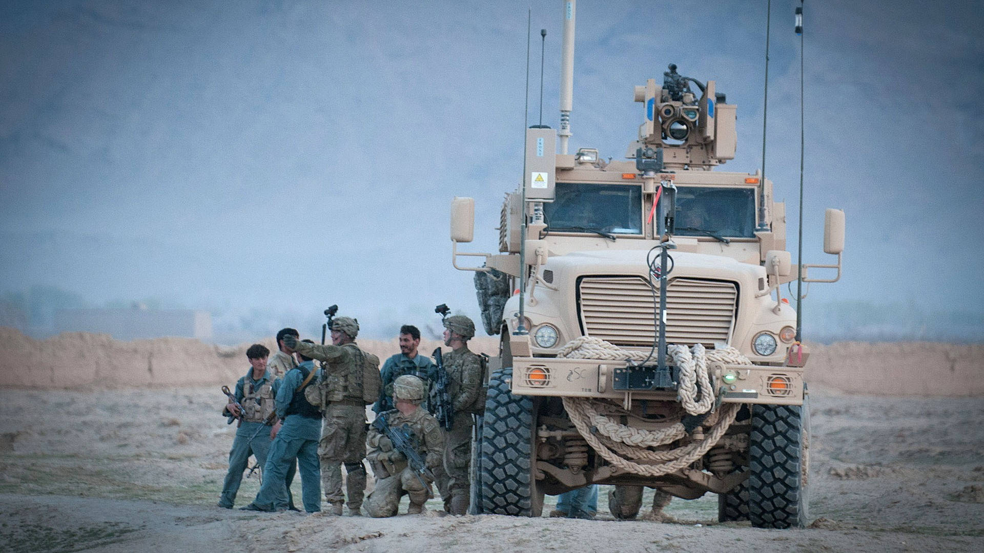 Afghanistan 桌布 壁紙 — 身穿迷彩裝備的士兵站在一輛大型棕褐色軍用卡車旁，背景是塵土飛揚的沙漠景觀，天空朦朧。
